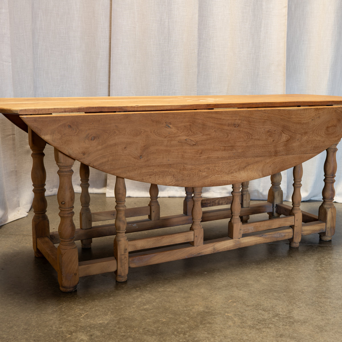 A rustic wooden drop-leaf table with turned spindle legs and a lower stretcher bar, shown against a light fabric backdrop and concrete floor.