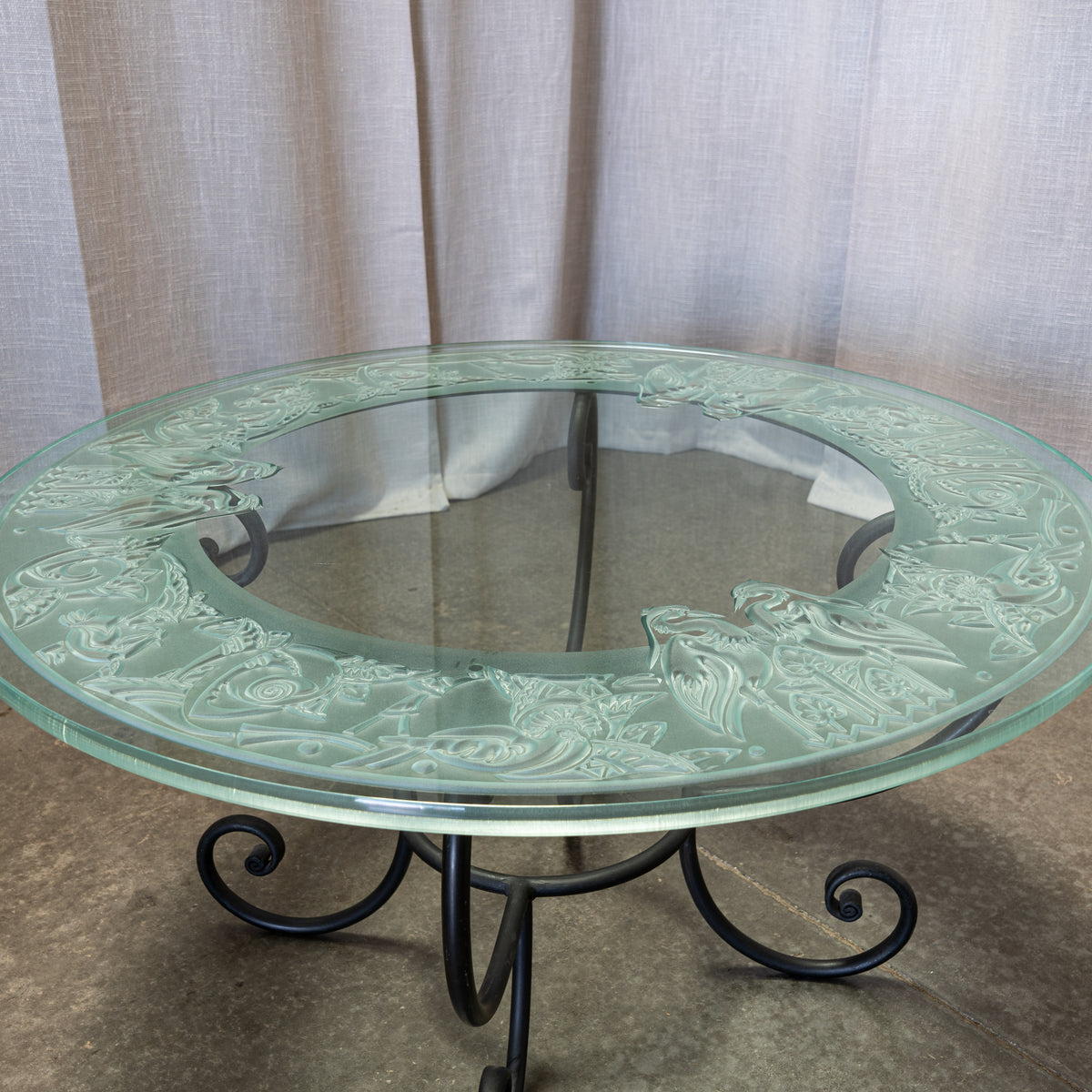 Round glass-top table with an ornate embossed floral border and a black metal scroll base, photographed indoors against draped fabric and a concrete floor.