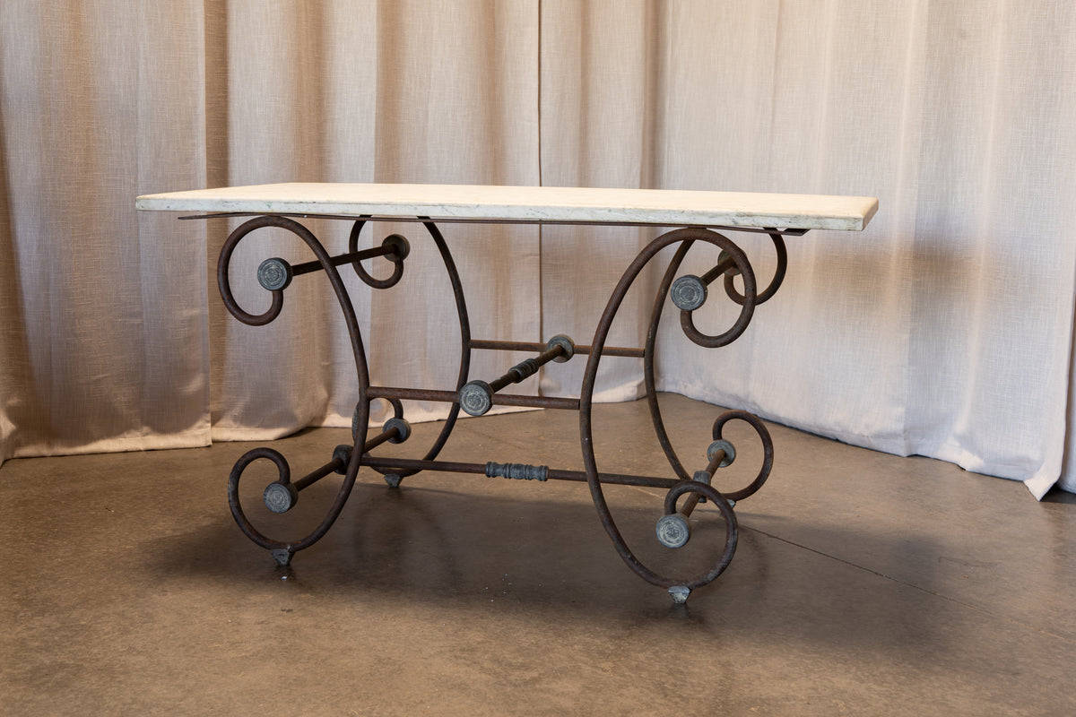 A vintage industrial-style console table featuring a rectangular white marble top and an ornate dark cast iron base with scrollwork and visible rivets, set against a draped beige fabric backdrop.