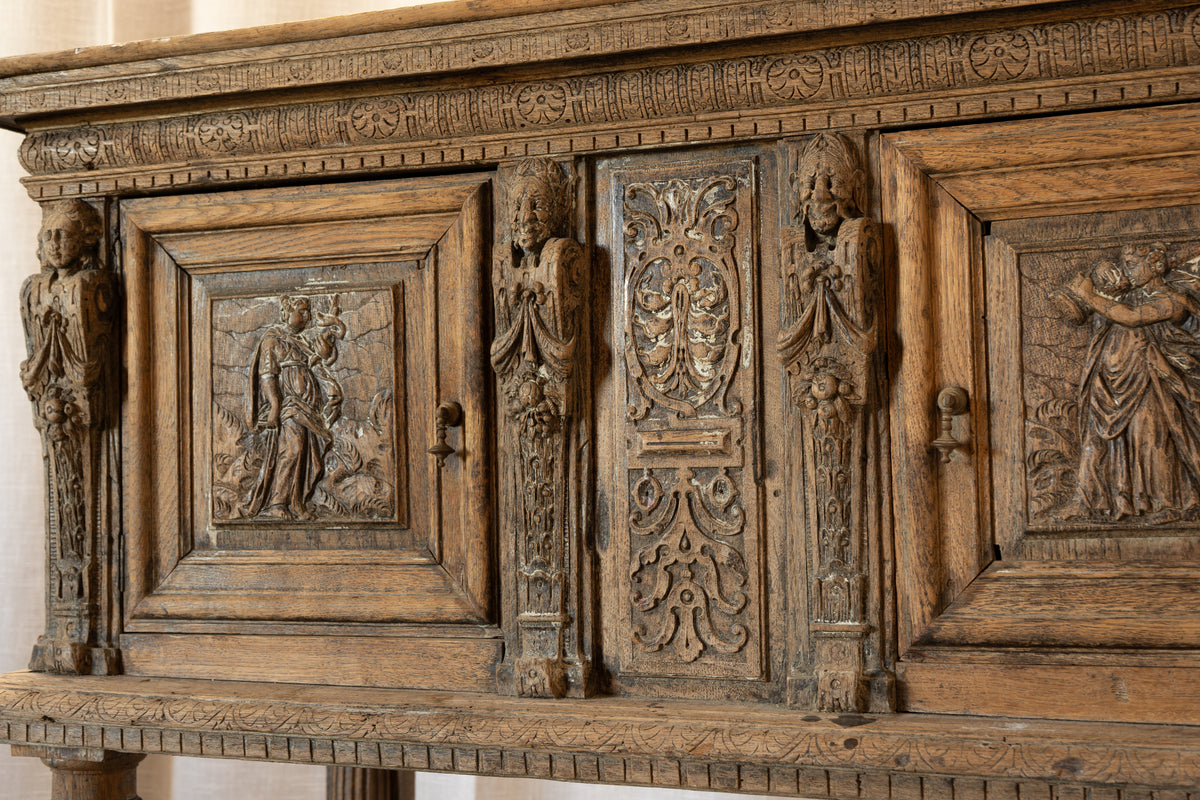 Close-up of an ornate carved wooden cabinet or sideboard with detailed relief panels, sculpted figures, and aged brown wood grain.