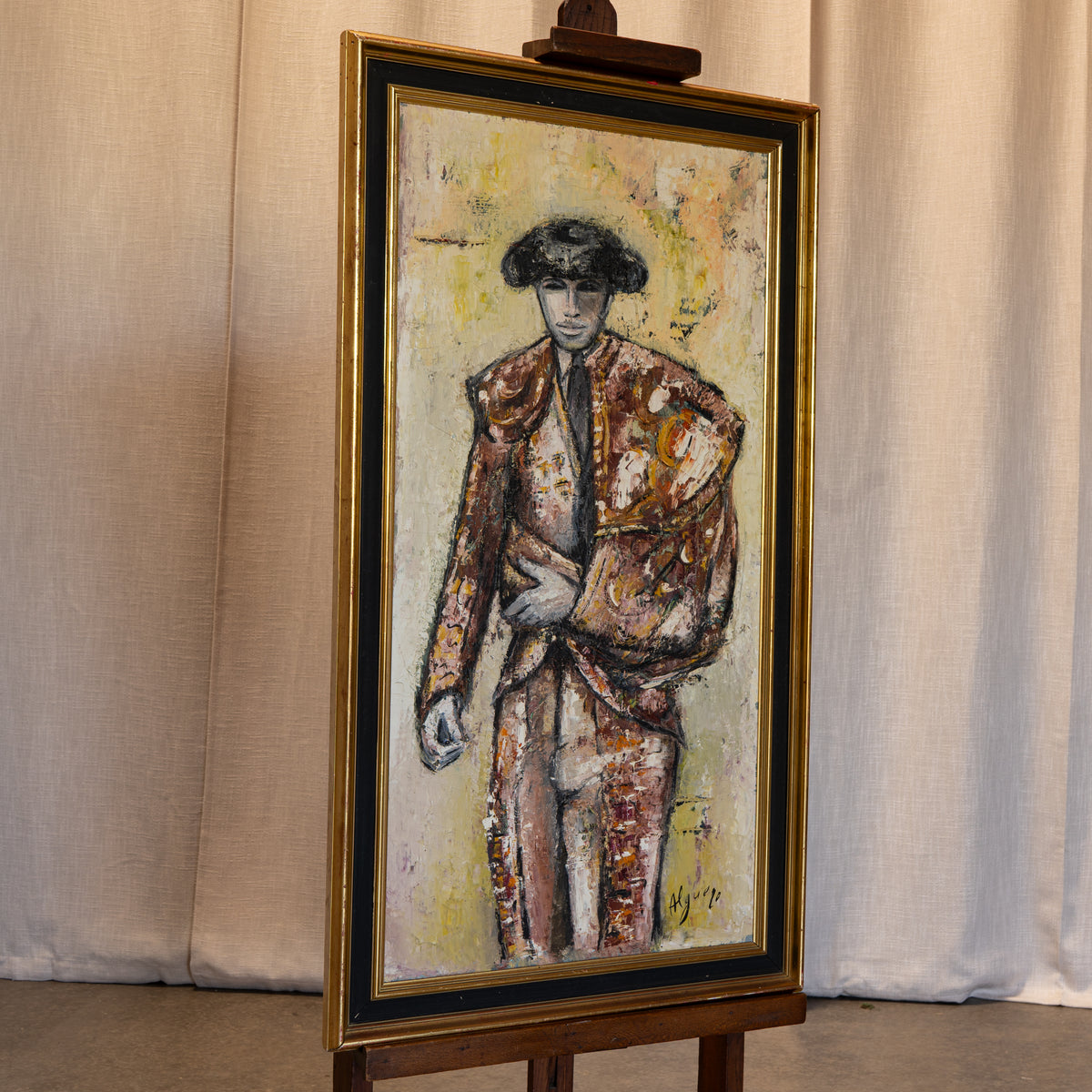 A framed vertical painting on an easel, depicting a stylized figure in a brown and gold suit against a pale yellow and beige background, set against a draped beige fabric backdrop.