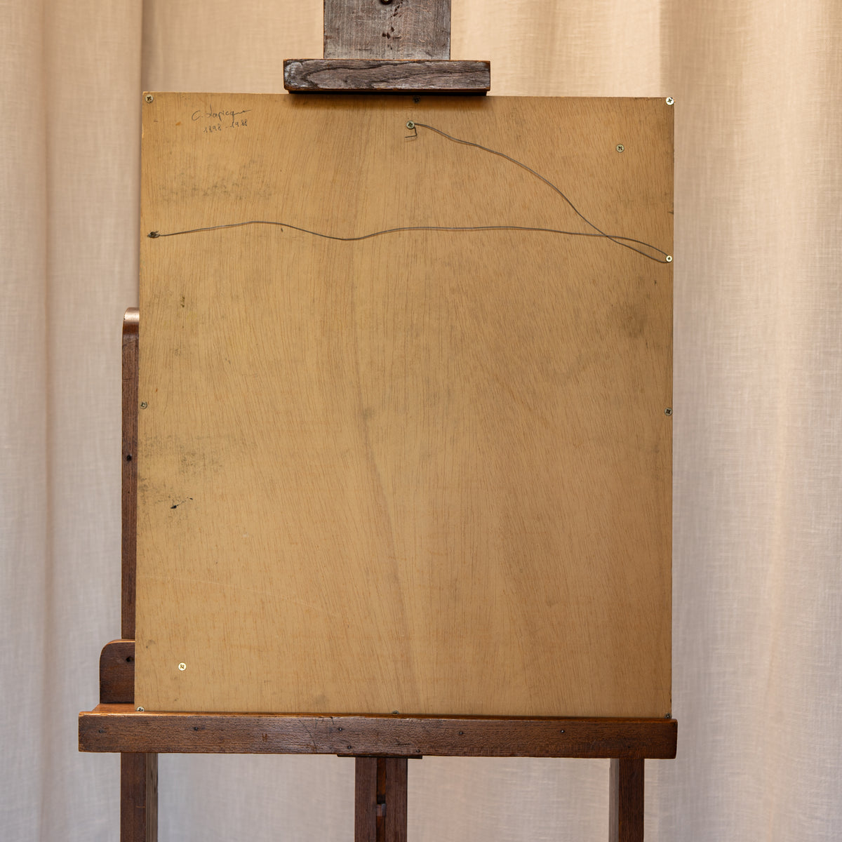 The back of a wooden art canvas or panel mounted on a dark wooden easel, with a simple curved line drawing and small handwritten markings near the top left, set against a neutral beige curtain background.