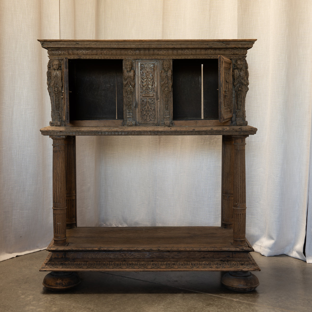 An antique carved wooden cabinet with two open upper compartments and a lower shelf, photographed against a draped off-white fabric backdrop.