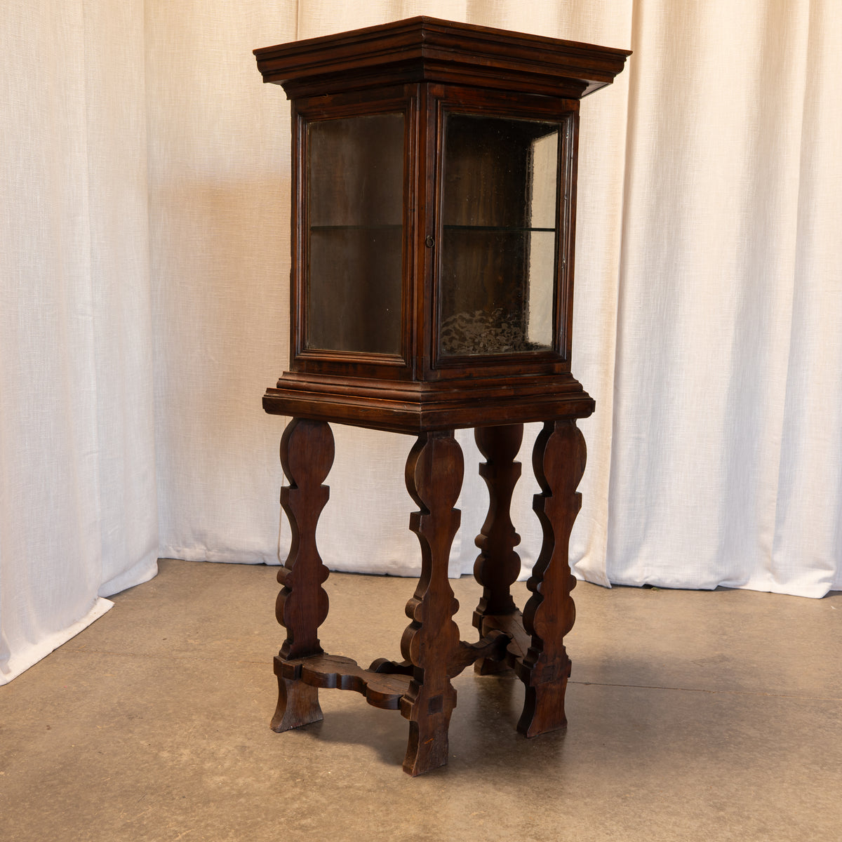 An antique dark wood display cabinet on a carved wooden stand, photographed against a draped white fabric backdrop.