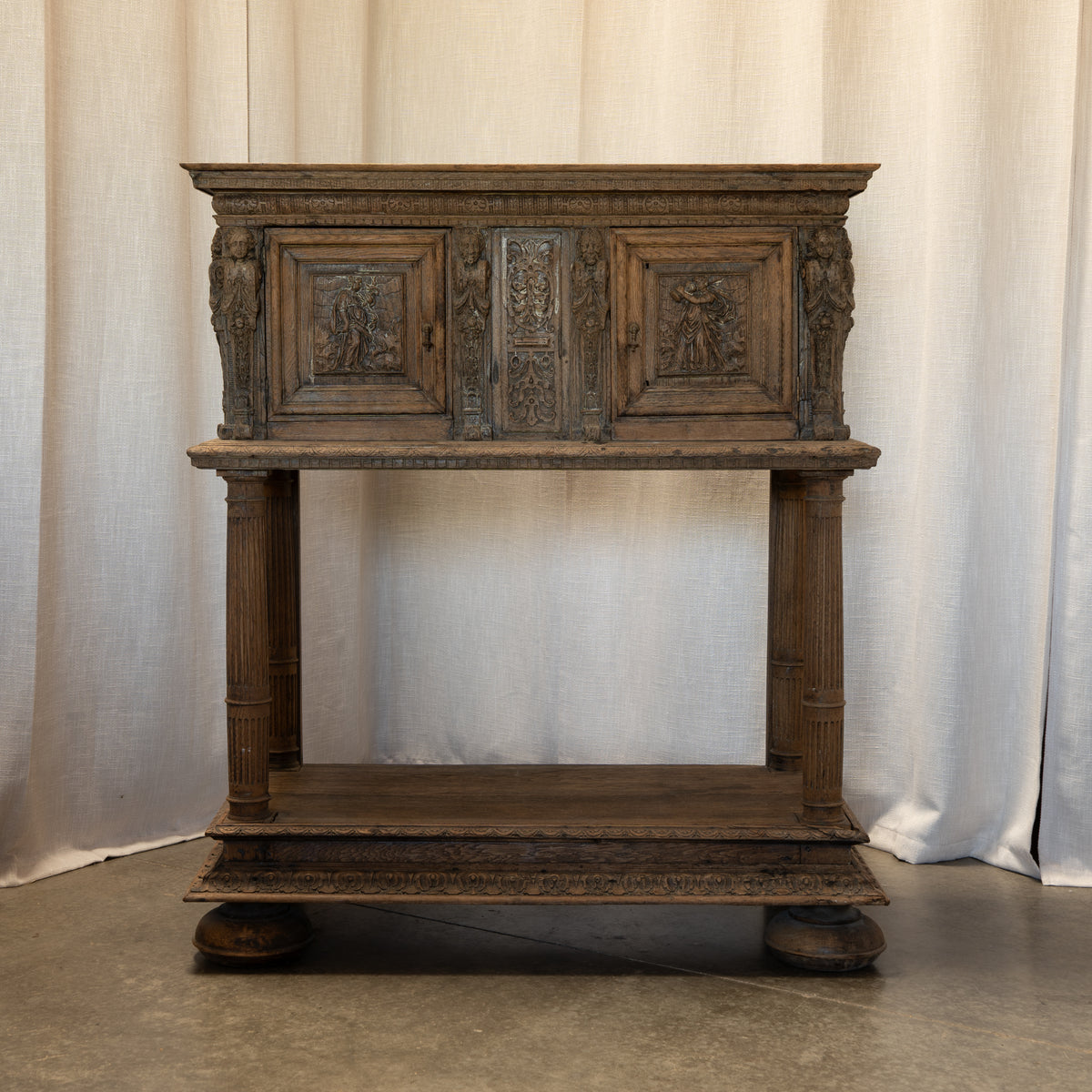 An ornate dark brown carved wooden console table with three cabinet doors, decorative panels, fluted legs, and a lower shelf, set against a draped beige fabric backdrop.