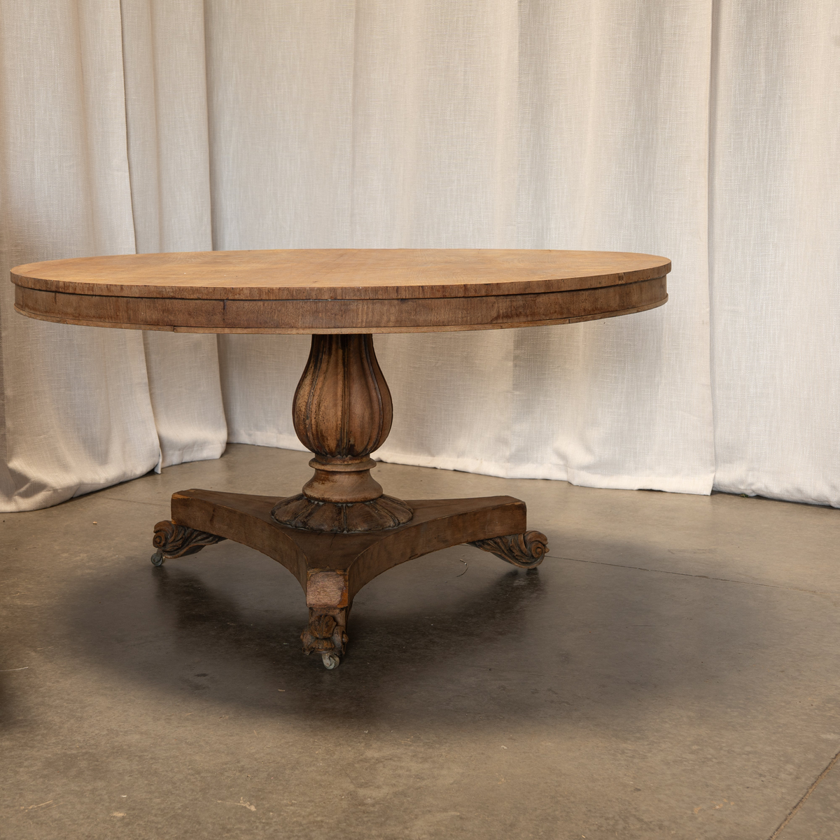 An antique round wooden pedestal table with ornate carved legs and casters, set against a neutral draped fabric backdrop.