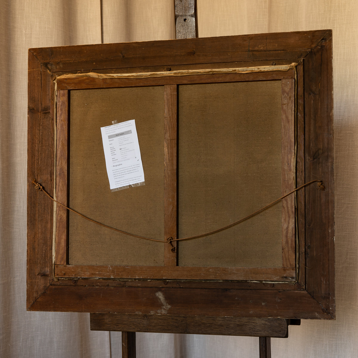 The back view of a rustic wooden picture frame mounted on an easel, showing a brown fabric backing with a white paper tag attached.