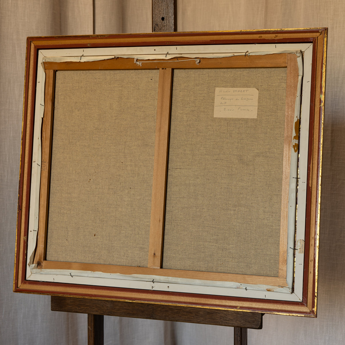The back side of a vintage framed canvas painting on an easel, showing the linen backing, wooden stretcher bars, and original gold and white painted frame.