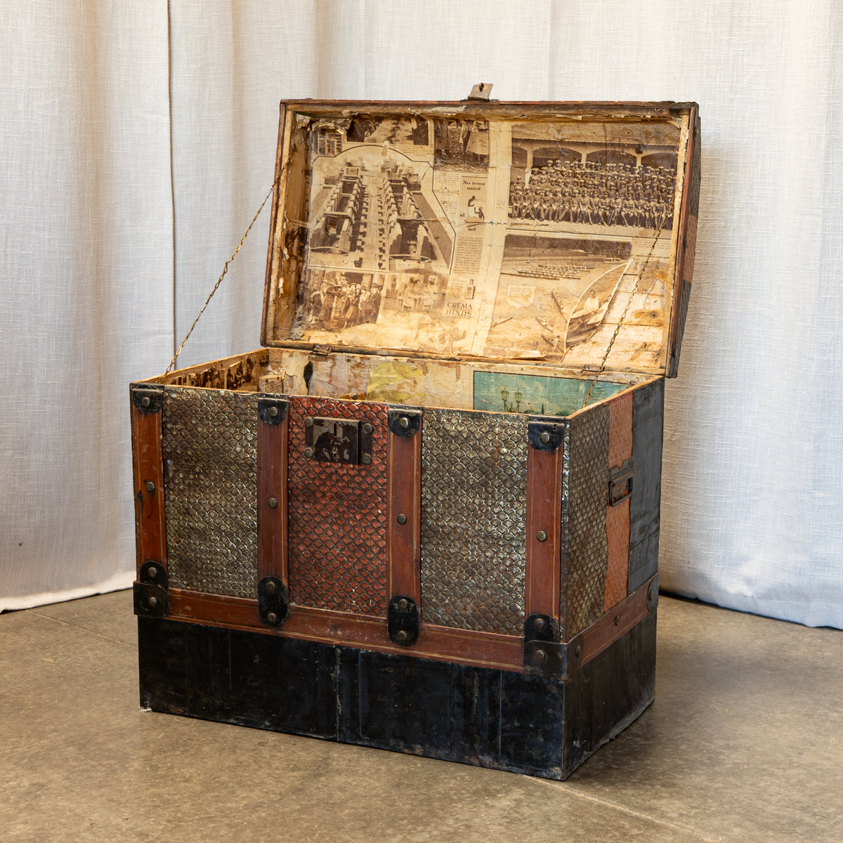An antique wooden trunk with a hinged lid, featuring a collage of vintage photographs on the inside lid, textured metal mesh panels, and a dark painted base, set against a neutral fabric backdrop.