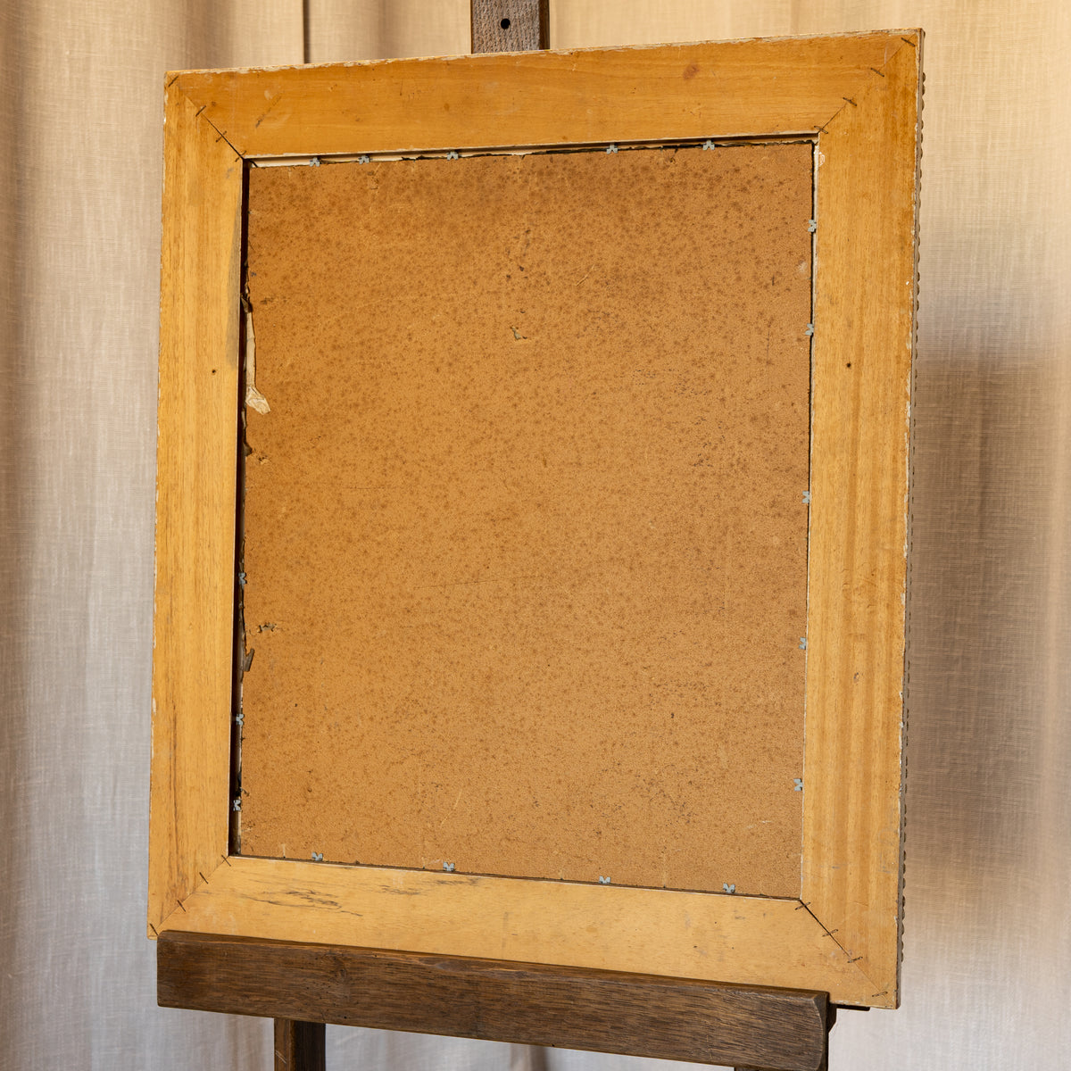 The back of a square wooden picture frame with a light natural wood border and a brown cork backing board, mounted on a dark wooden easel against a beige linen background.
