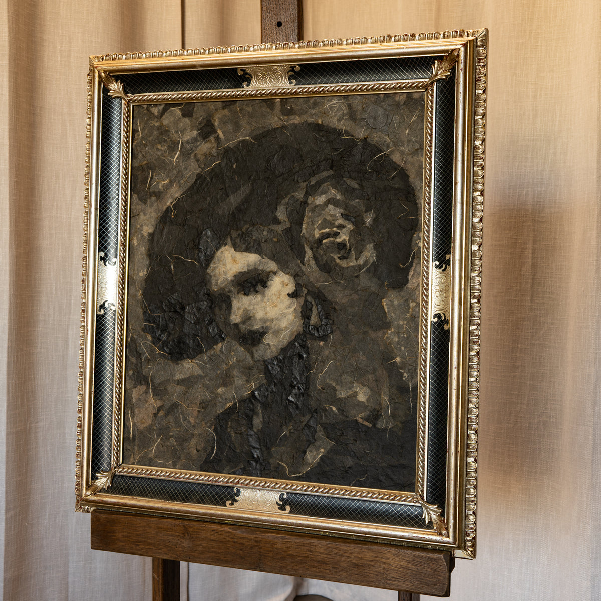 A framed portrait painting of two figures in a dark, textured style, displayed on a wooden easel against a beige curtain background. The frame is ornate and gold-toned with black textured accents.