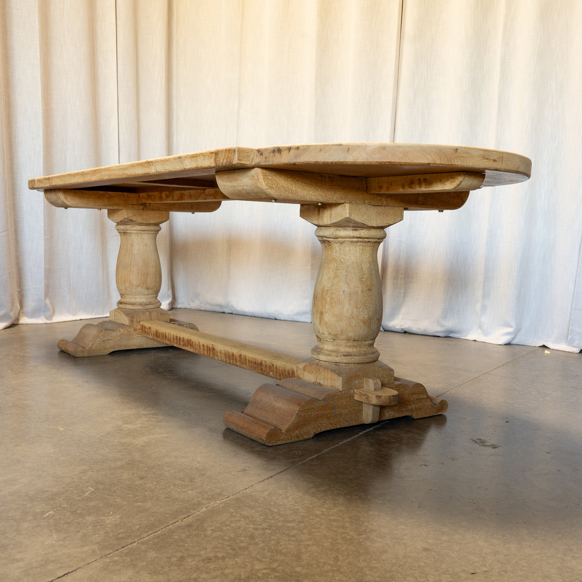 A rustic light oak trestle dining table with carved turned legs and a stretcher base, set against a draped cream fabric backdrop.