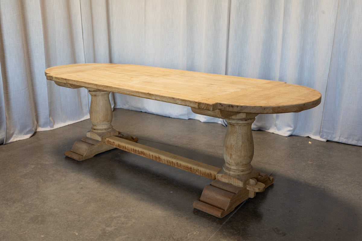 An oval wooden dining table with a light natural wood top and a weathered, rustic gray-brown trestle base, photographed against a light blue curtain backdrop.