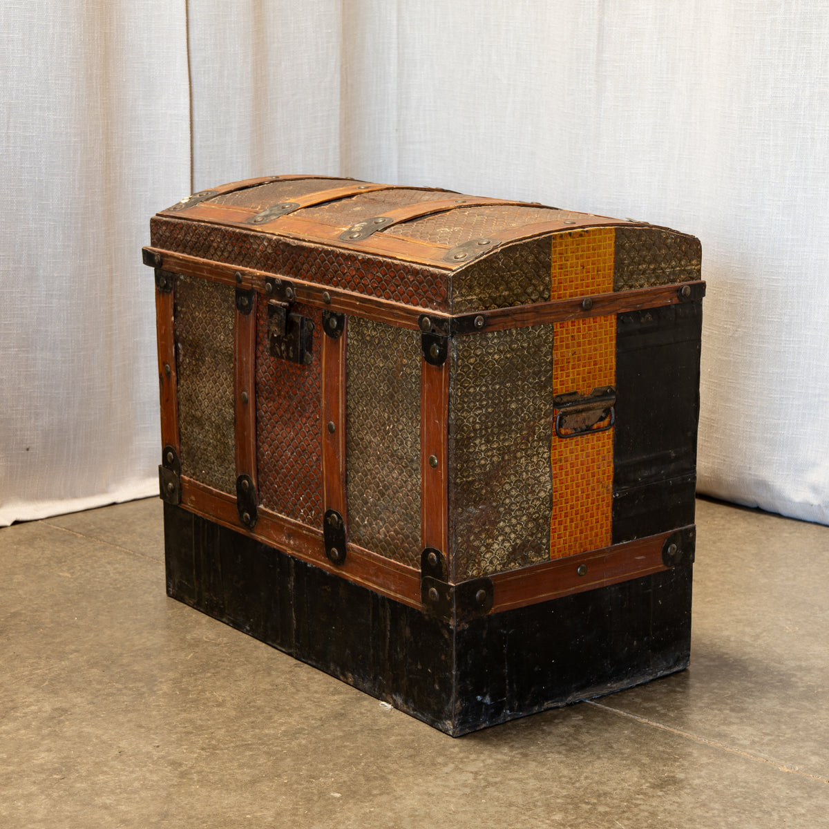 A vintage wooden trunk chest with woven rattan panels, dark metal corner protectors, and a black-painted base, photographed against a draped fabric backdrop.