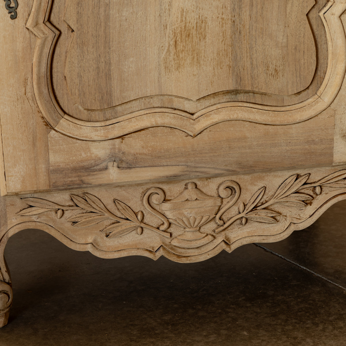 Close-up of a carved wooden cabinet or chest with ornate floral scrollwork and a distressed light tan finish, showing part of the upper panel and lower decorative apron.