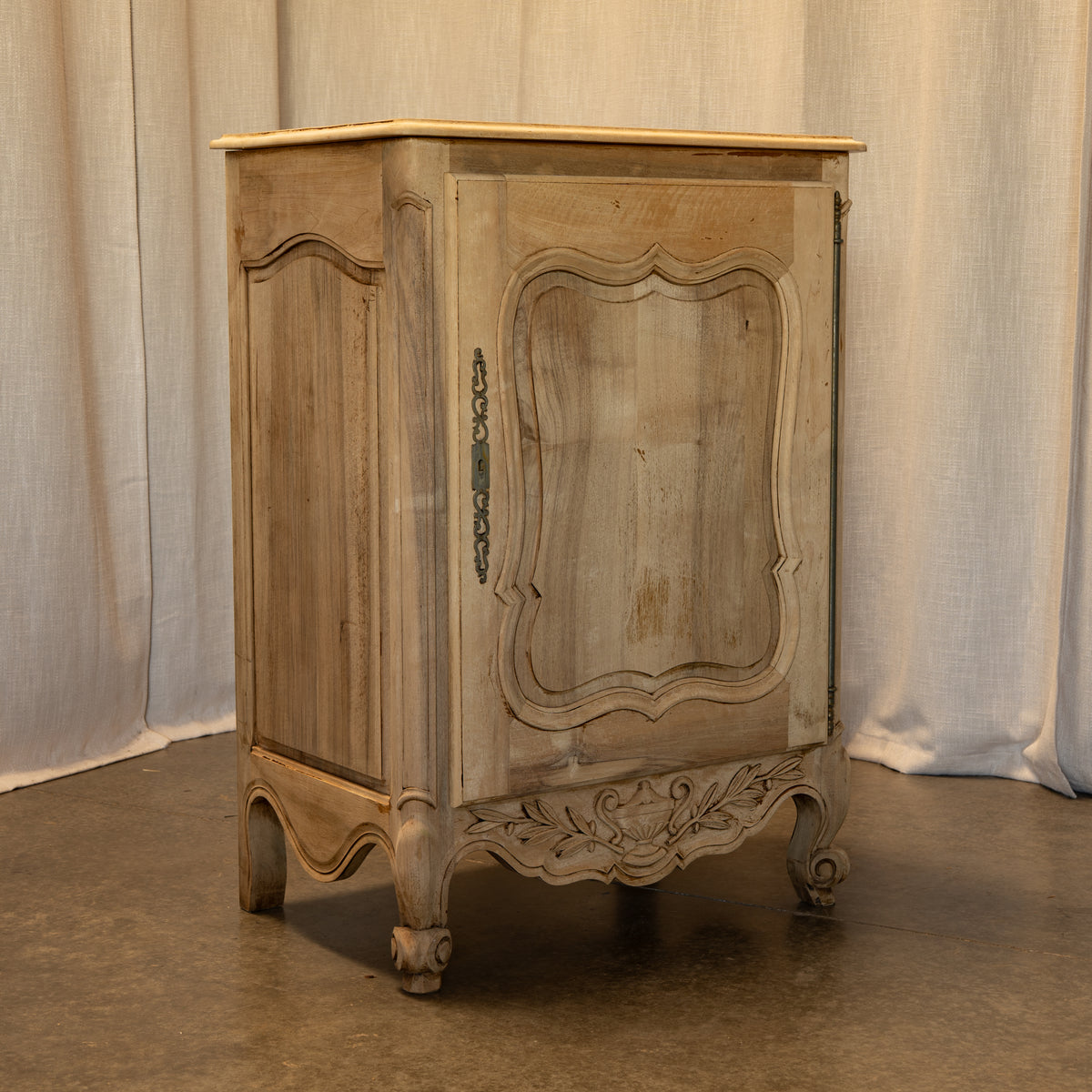 A distressed, vintage-style French provincial nightstand or cabinet with carved details, featuring a light beige wood top and a weathered grayish-beige painted finish, set against a neutral draped fabric backdrop.