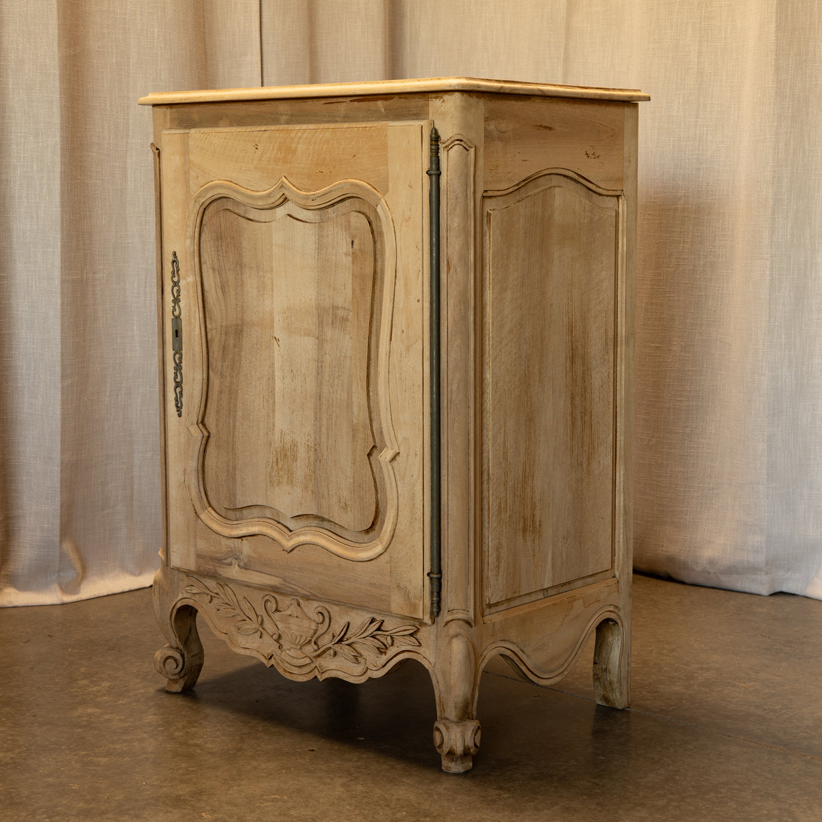 A distressed vintage-style wooden cabinet with ornate carved details and cabriole legs, finished in a light beige/gray wash, set against a neutral draped fabric backdrop.