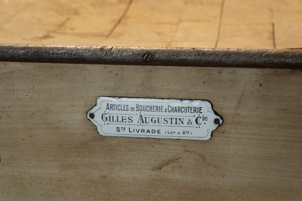 A close-up of a vintage metal sign reading 'Articles de Boucherie & Charcuterie Gilles Augustin & Cie' mounted on a light wood surface.