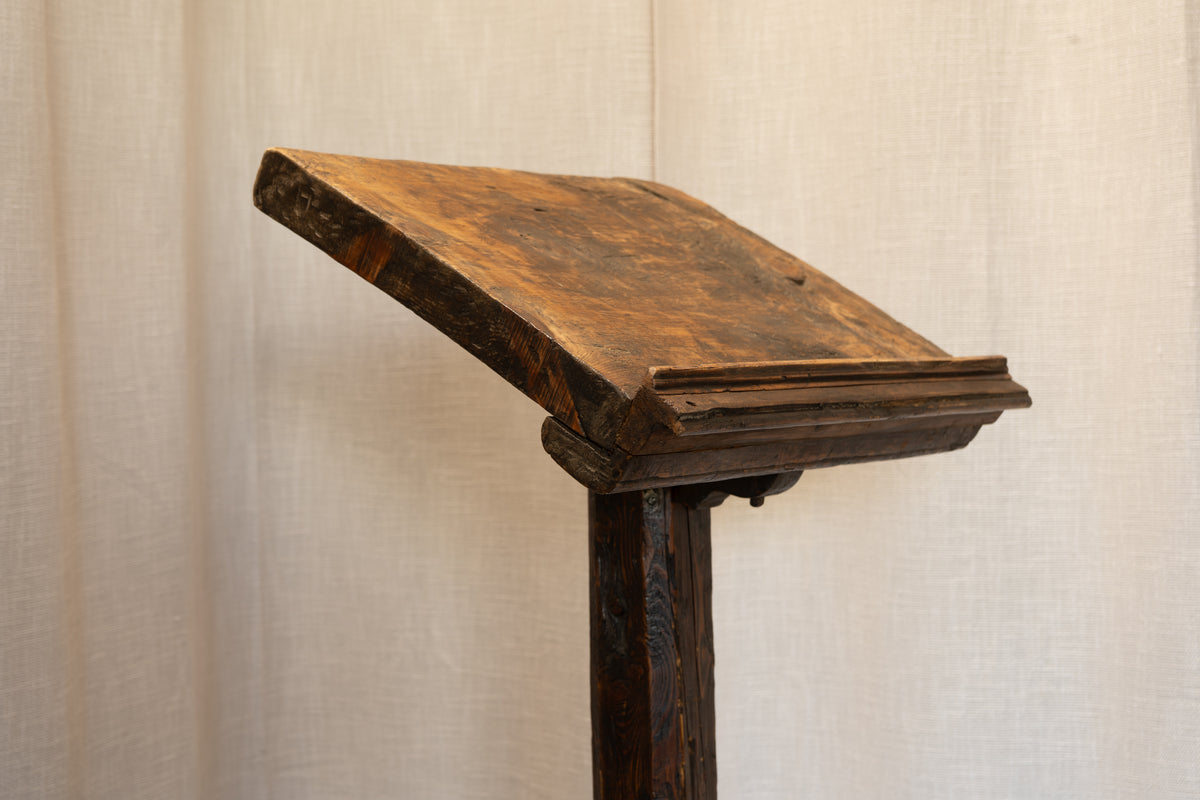 A rustic dark brown wooden lectern or stand with a slanted reading surface, shown against a neutral beige fabric backdrop.