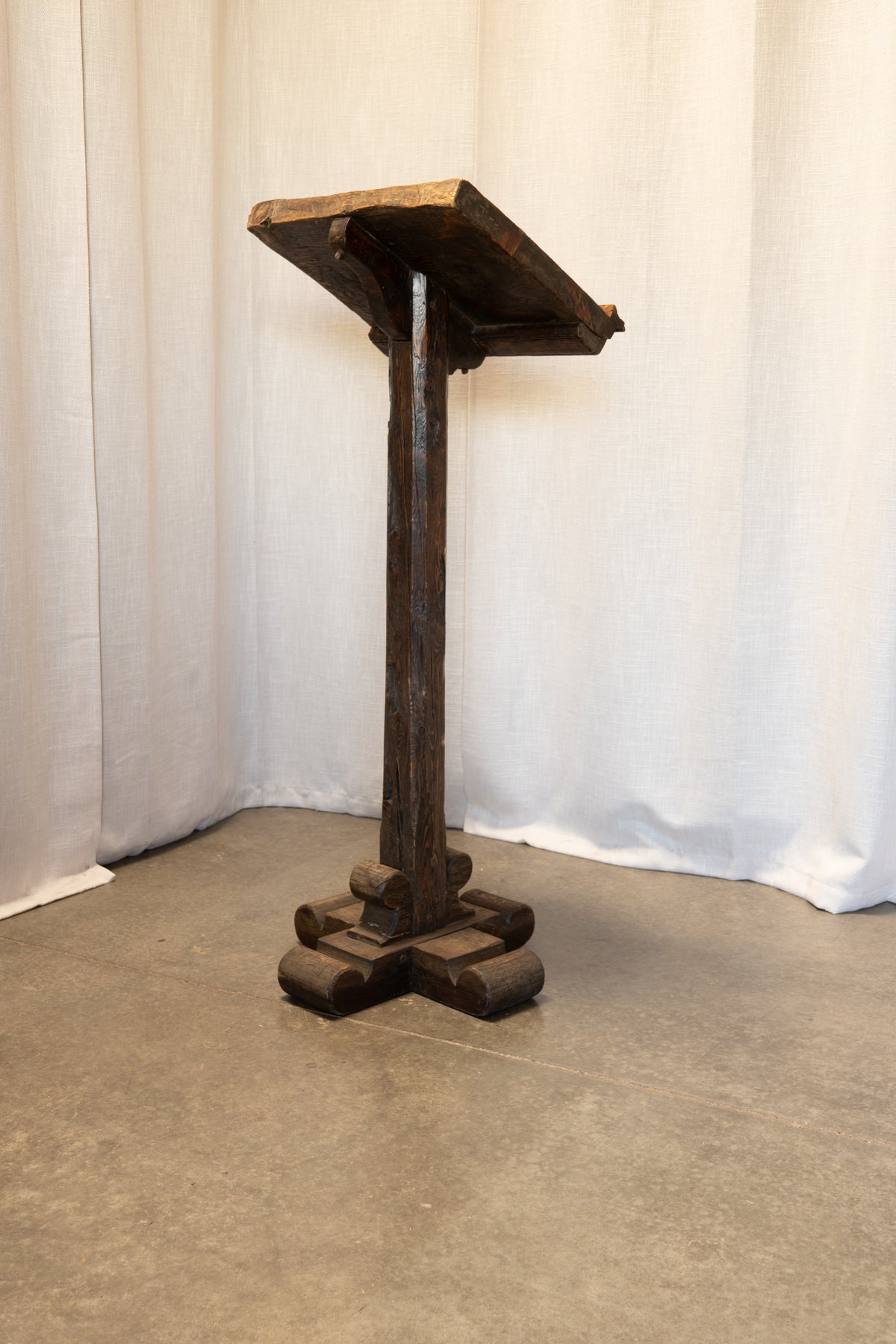 A dark rustic wooden lectern or stand with a slanted top platform, single central post, and blocky tiered base, photographed against a white draped backdrop on a gray floor.