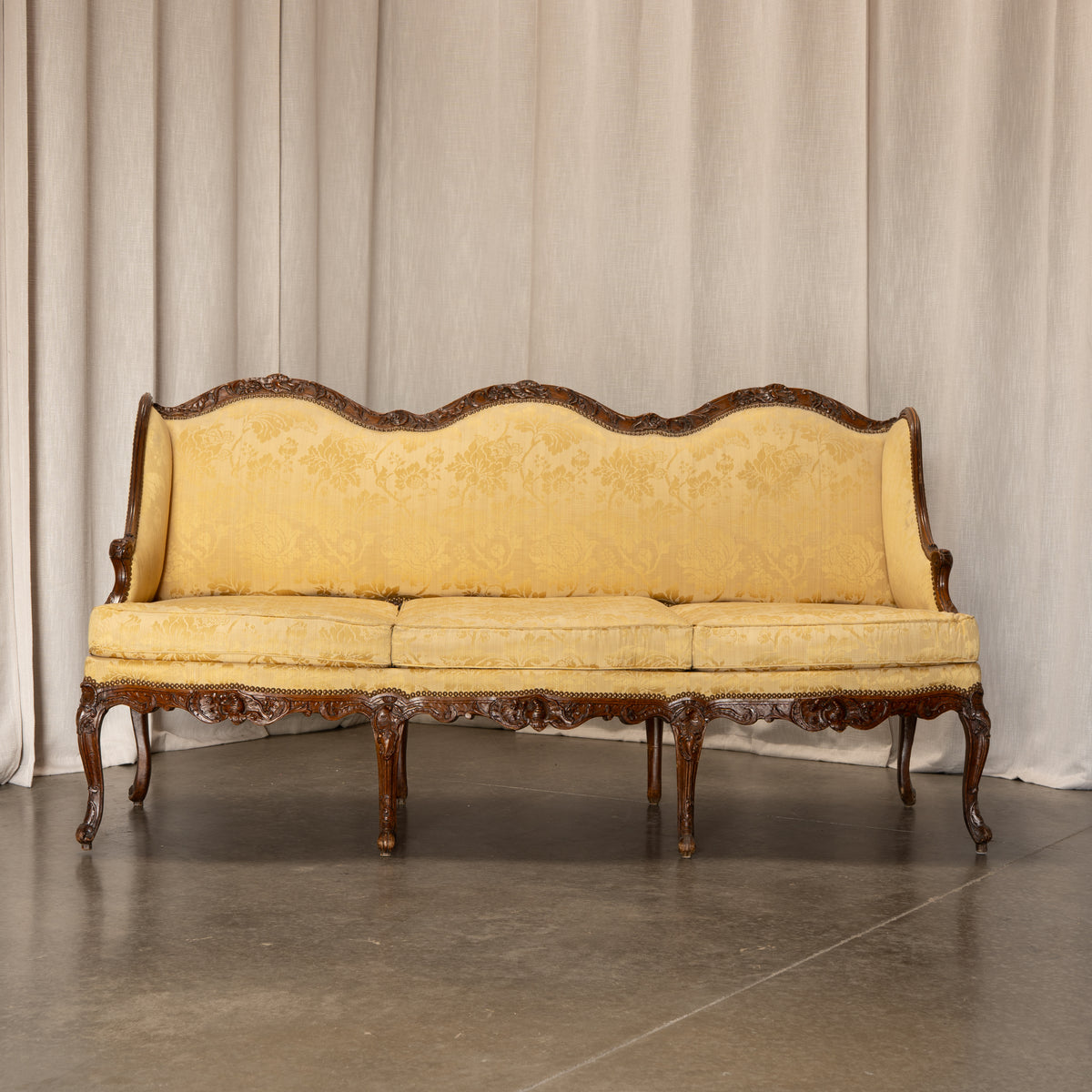An ornate vintage-style sofa with carved dark wood arms and legs, upholstered in a textured pale yellow fabric, set against a neutral beige curtain backdrop.