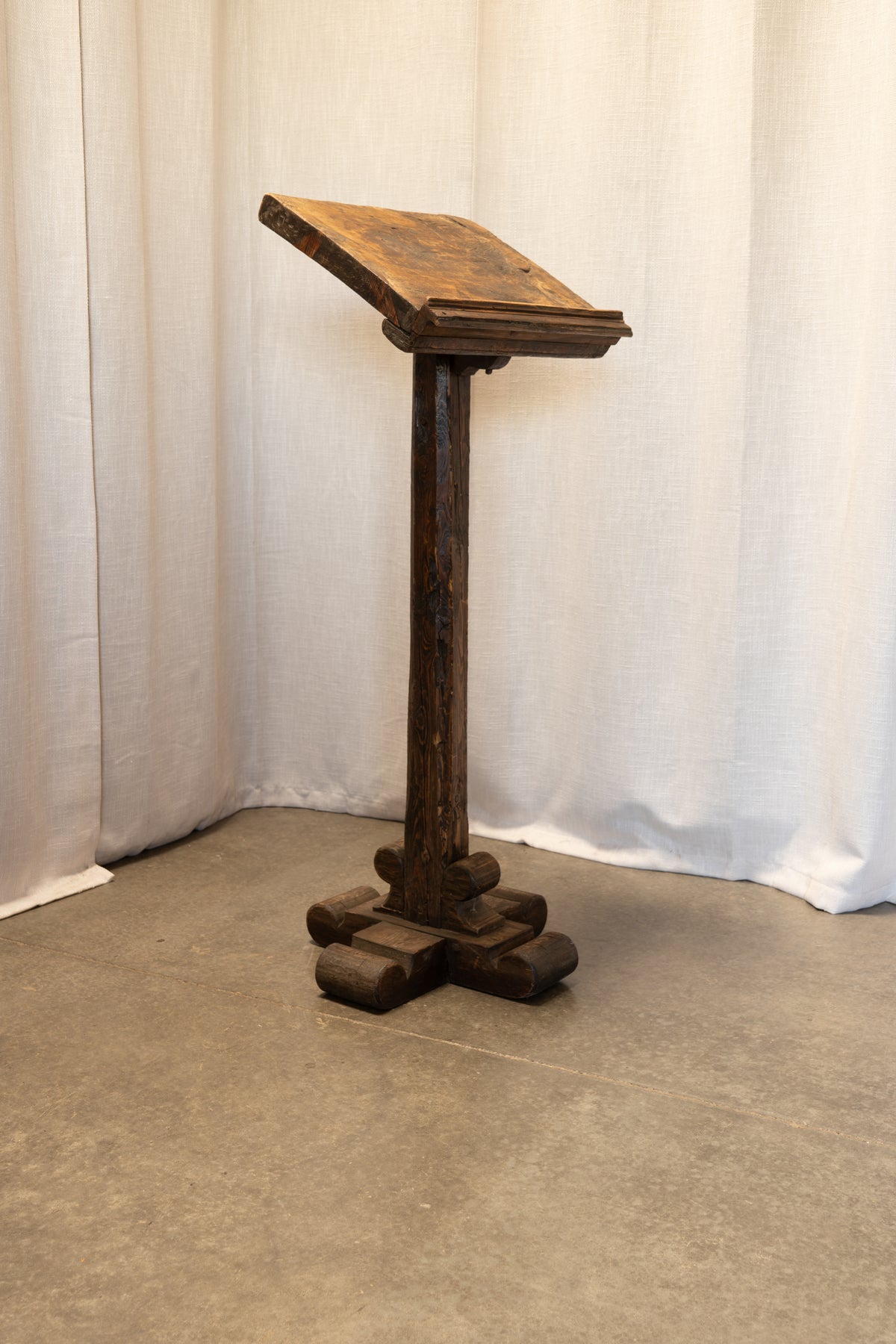 A dark brown carved wooden lectern or stand with a slanted reading surface, photographed indoors against a draped white fabric backdrop on a concrete floor.