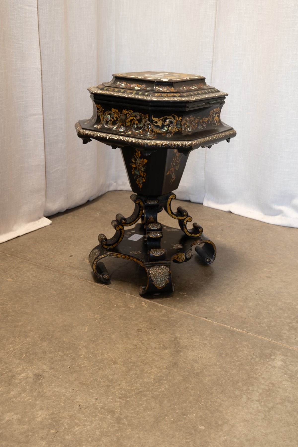 An ornate black and gold decorative pedestal table or stool with carved floral detailing, shown on a concrete floor against a draped white fabric backdrop.