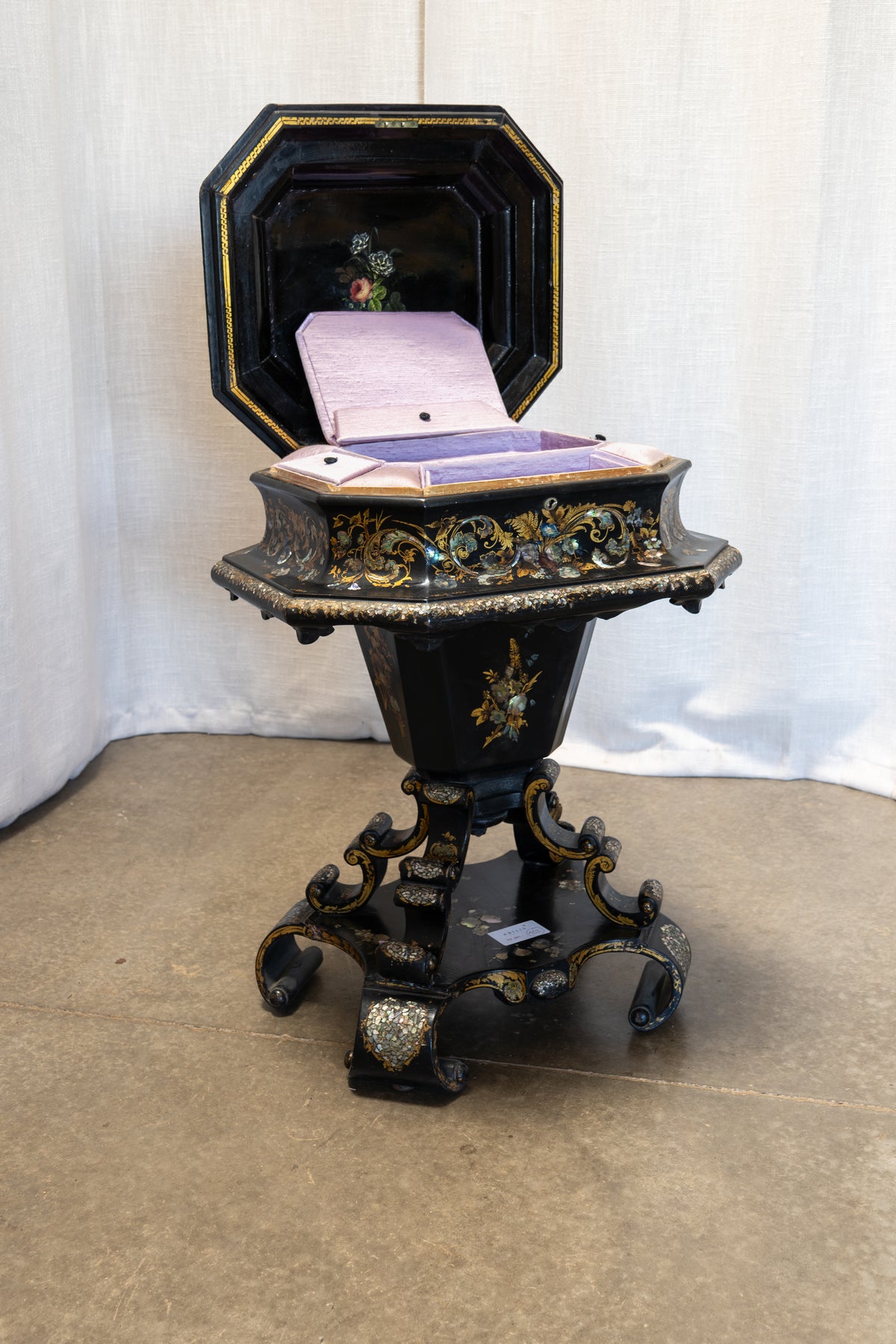 An ornate black octagonal pedestal table with gold and silver decorative accents, featuring an open lid that reveals a light purple velvet-lined interior with a small floral painting.