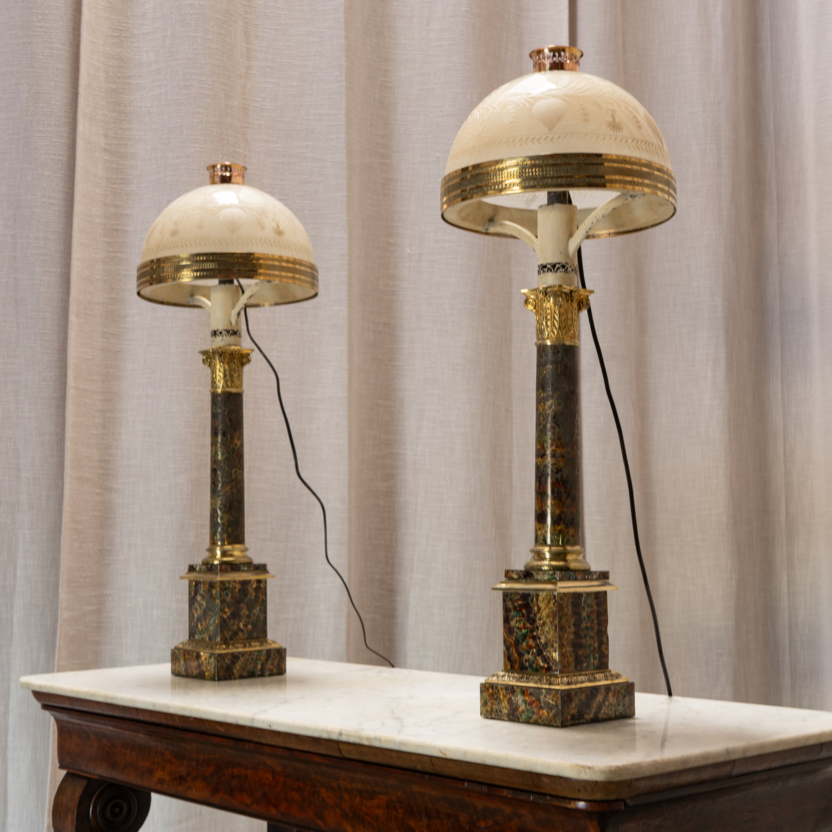 Two matching ornate table lamps with cream dome shades, gold trim, and dark marbled bases, displayed on a white marble-topped table against beige curtains.