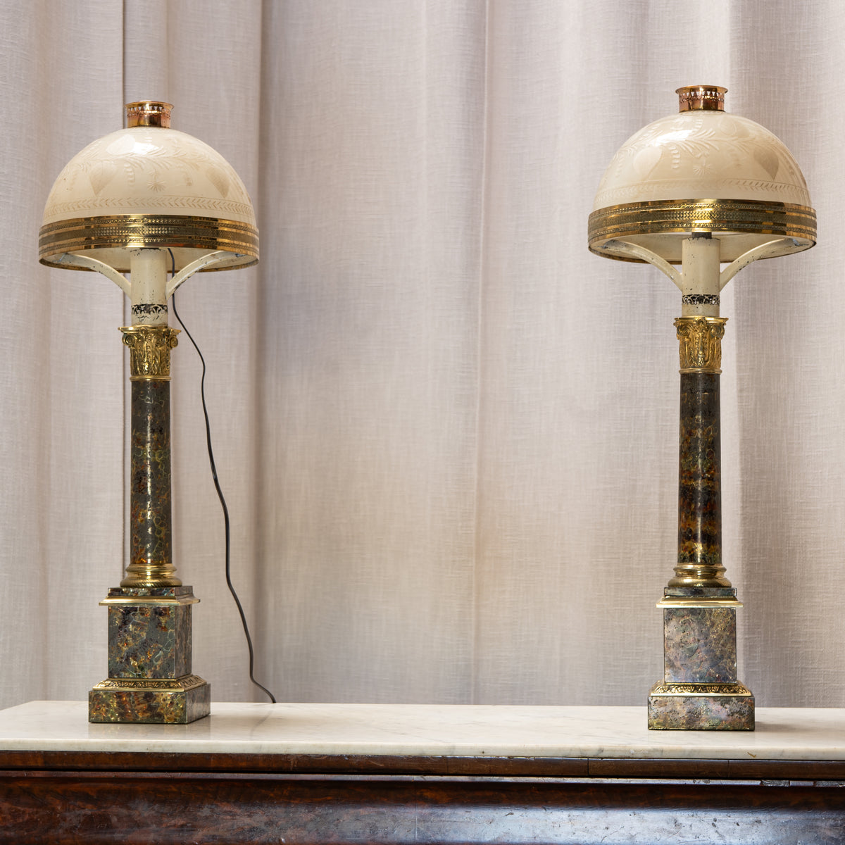 A pair of ornate table lamps with cream-colored dome shades, gold trim, and dark marble columns with gold accents, displayed on a marble mantelpiece in front of a beige curtain.