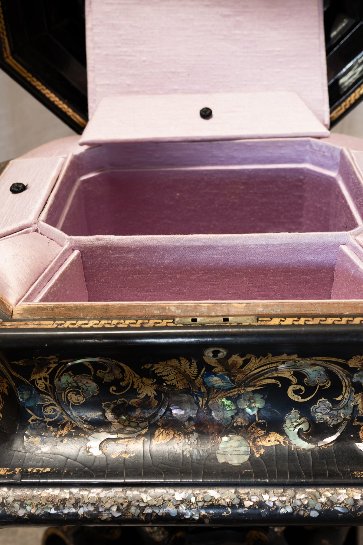 An ornate black lacquered jewelry box with gold and iridescent floral detailing, shown open to reveal a light pink satin-lined interior.