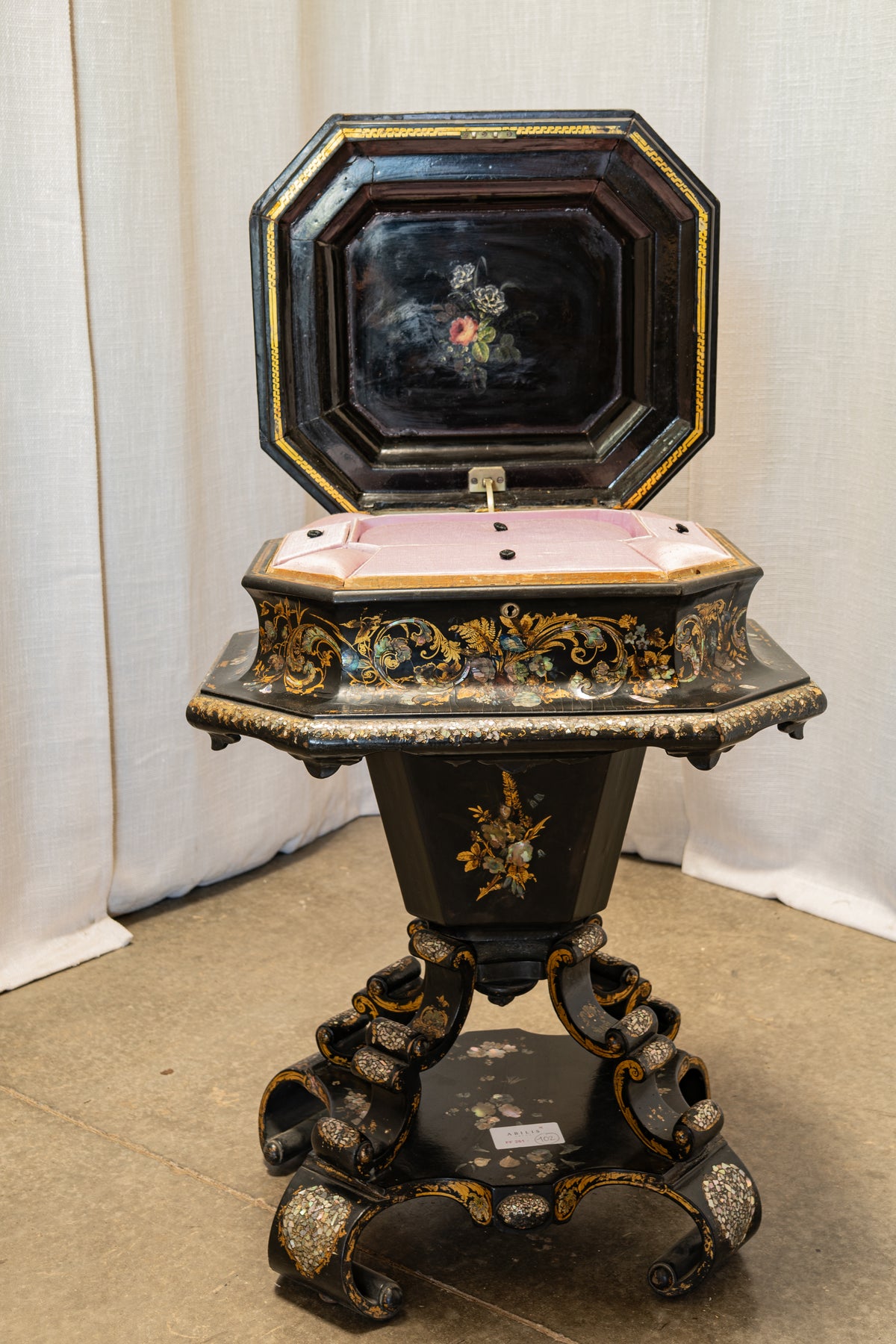 An ornate octagonal black wooden music box table with gold and silver gilding, a floral painted interior lid, and a pink fabric-covered turntable, photographed indoors against a white curtain backdrop.
