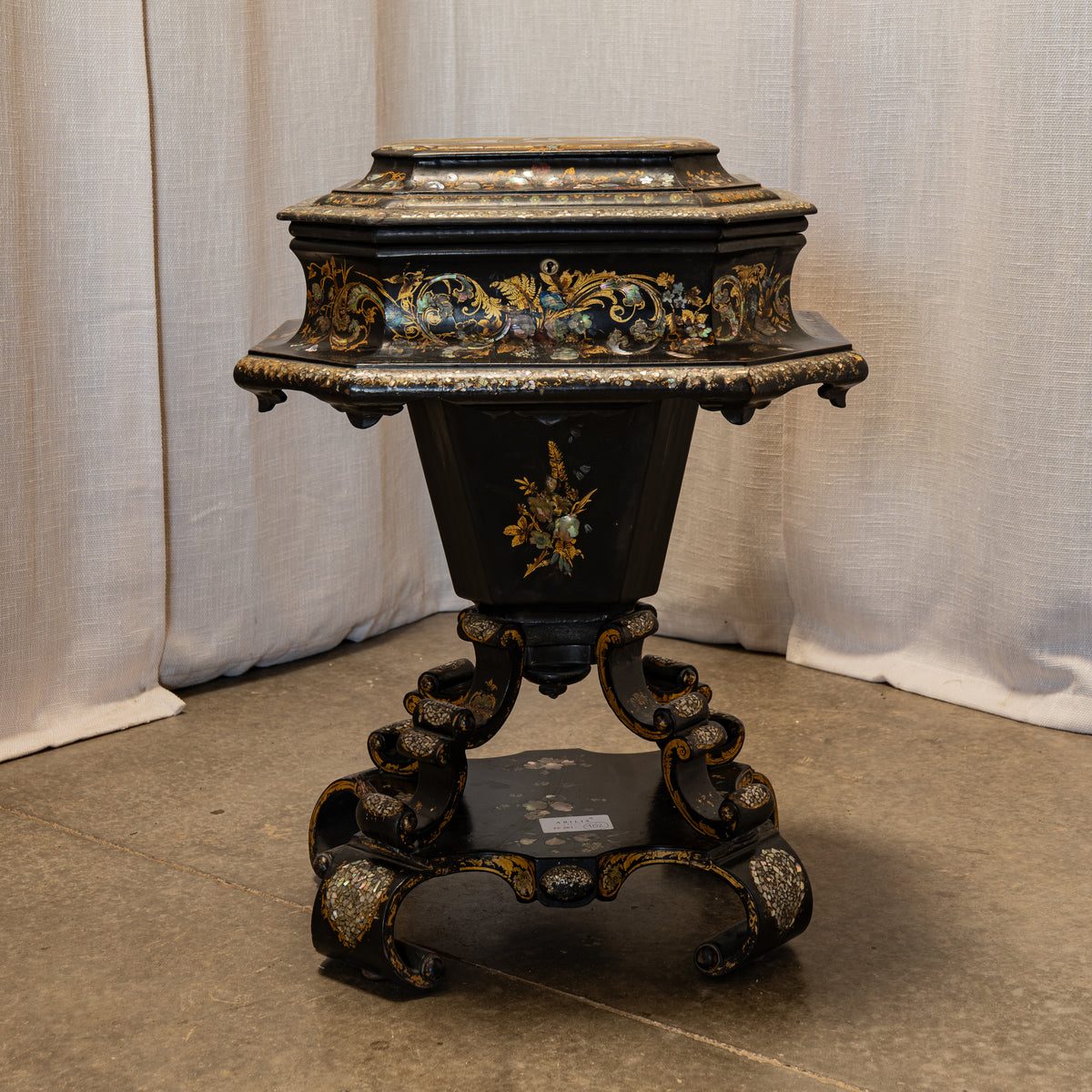 An ornate black octagonal pedestal table with intricate gold and multicolor floral inlay detailing, set against a neutral draped fabric backdrop.