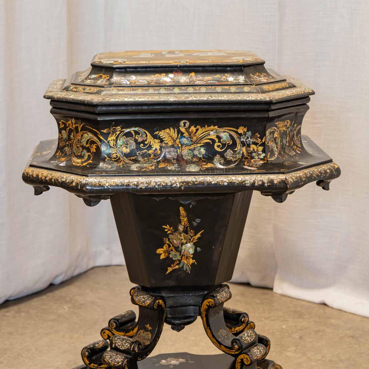 An ornate black octagonal pedestal table or stand with intricate gold, blue, and green floral inlay detailing, set against a draped white fabric backdrop.