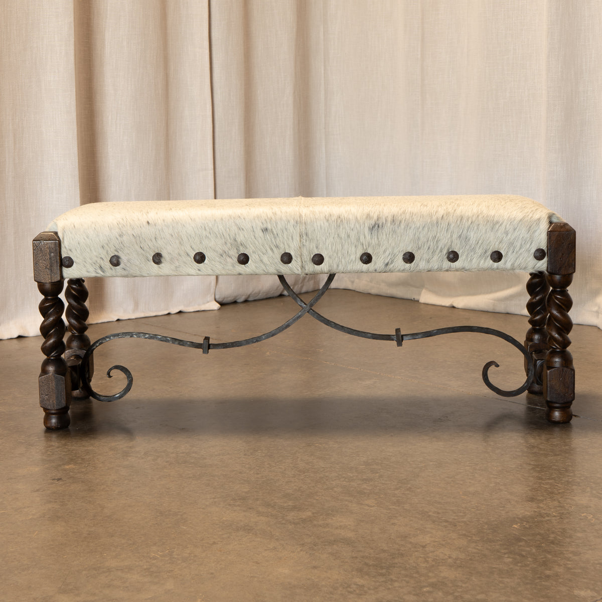 A rustic bench with a light-colored cowhide upholstered seat, dark metal nailhead trim, and dark brown twisted wooden legs on a wrought iron base, set against a neutral draped curtain backdrop.