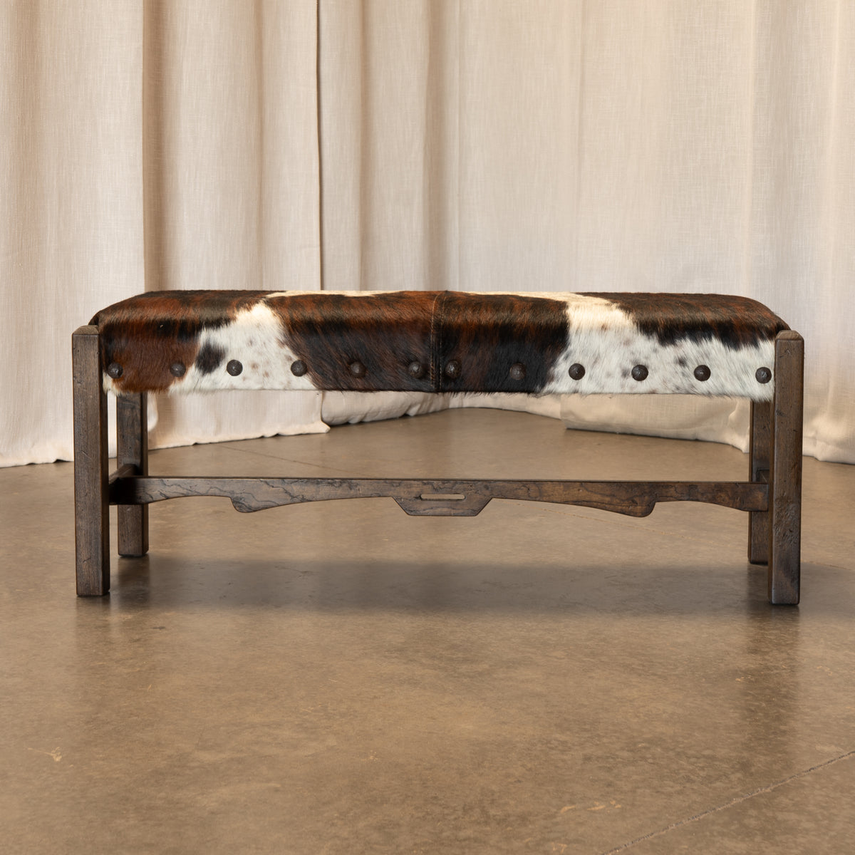 A rustic wooden bench with a brown, black, and white cowhide upholstered seat, featuring metal nailhead trim and a dark wood frame, set against a neutral beige curtain backdrop.
