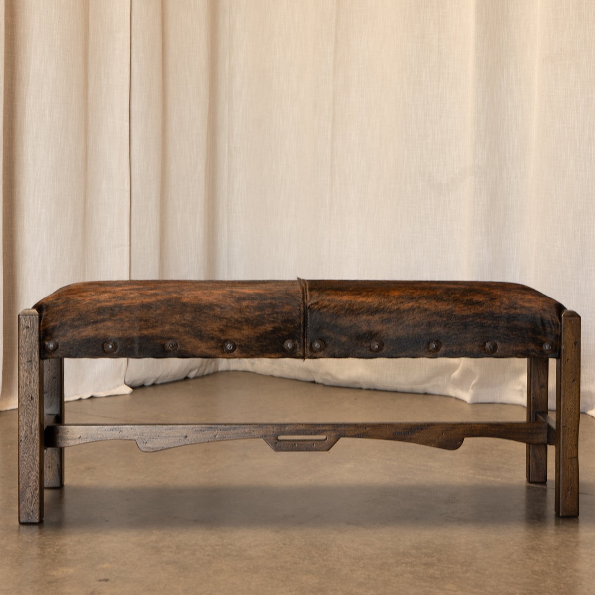 A rustic wooden bench with a dark brown cowhide upholstered seat, featuring metal nailhead trim and a lower wooden stretcher bar, set against a neutral beige curtain backdrop.