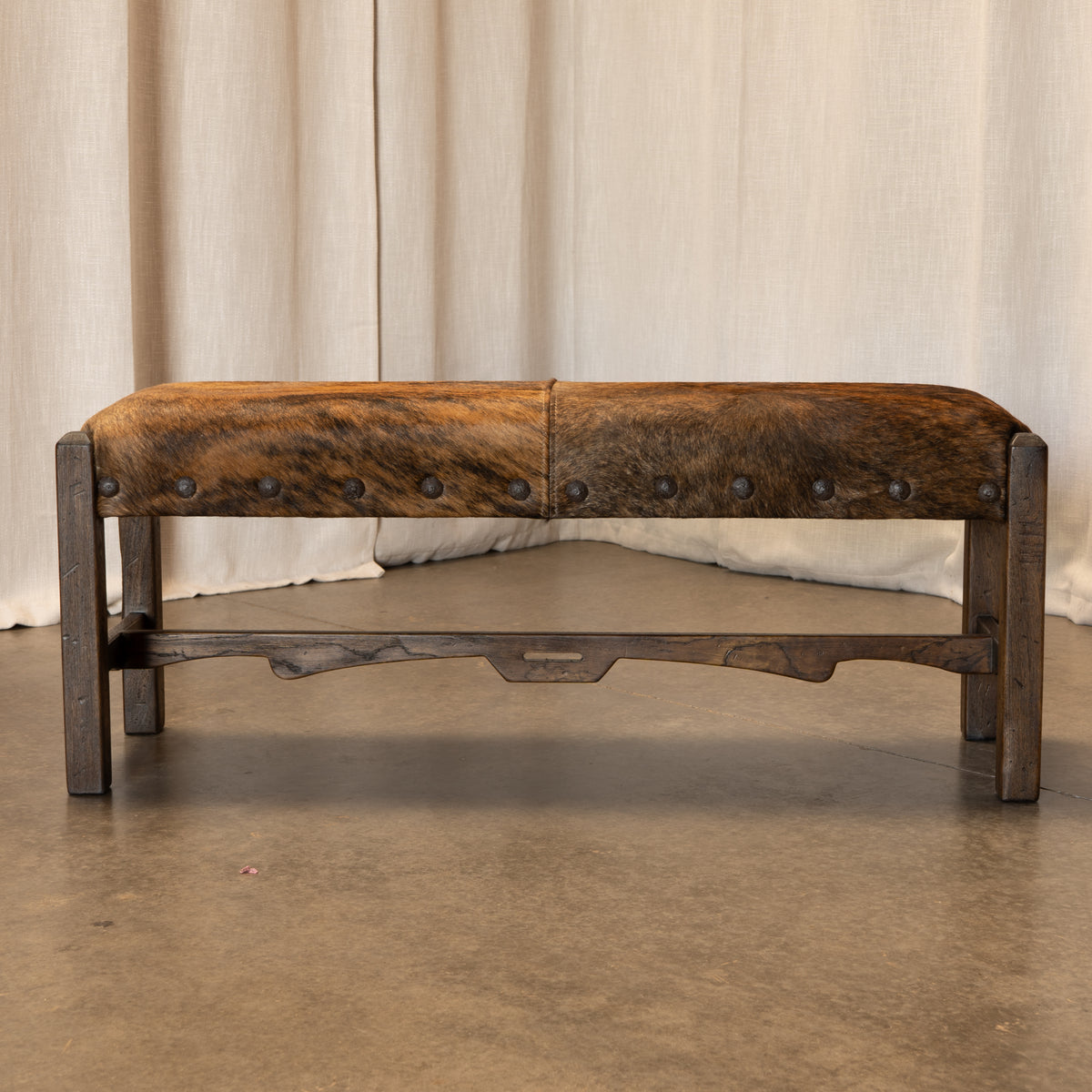 A rustic wooden bench with a cowhide upholstered top and dark stained wood legs, set against a neutral beige curtain backdrop.