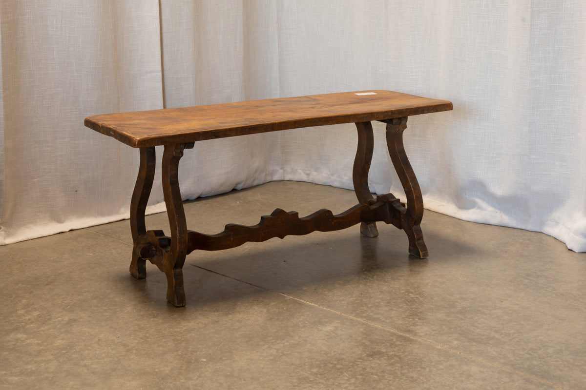 A rustic wooden bench with a rectangular top and curved legs, set against a draped white fabric backdrop on a concrete floor.