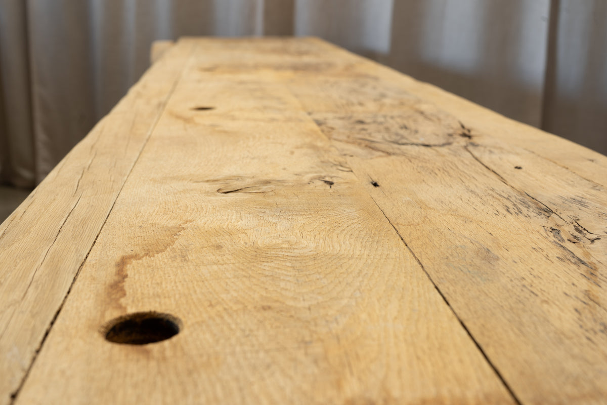 A close-up view of a rustic light oak wood tabletop with visible grain, knots, and natural imperfections, photographed indoors against a softly blurred neutral background.