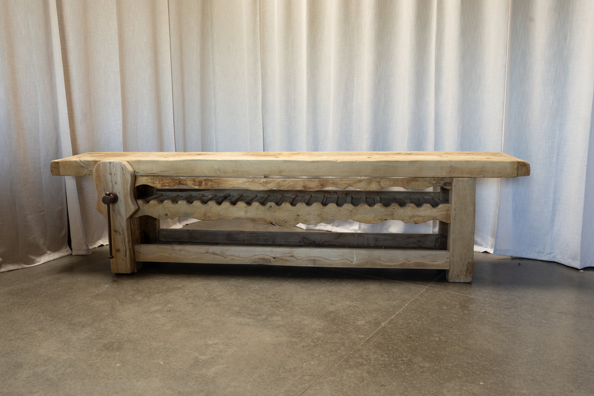 A rustic wooden bench with a thick natural wood top and legs, featuring a scalloped decorative apron and a dark metal support beam underneath, set against a white curtain backdrop.
