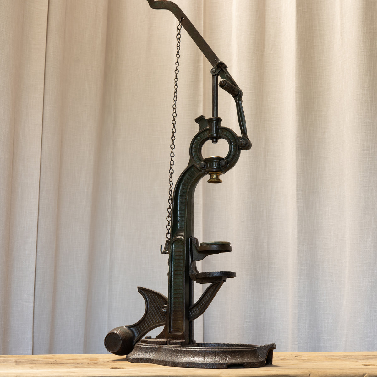 An antique-style black cast iron mechanical device with a chain, lever, and circular base, displayed on a light wood surface in front of a beige curtain backdrop.
