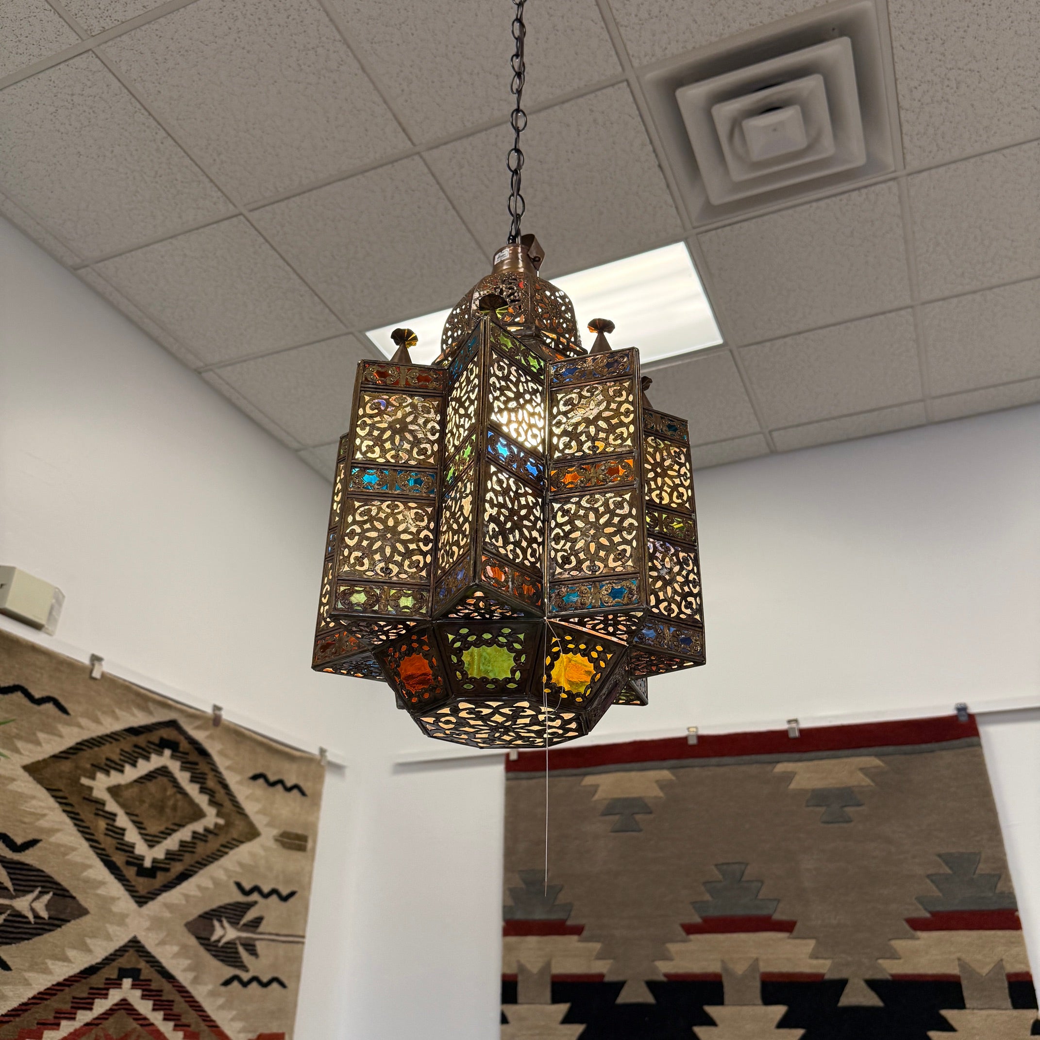 A hanging Moroccan-style metal lantern with intricate cutout patterns and colorful stained glass panels, suspended from a chain in a showroom with rugs and a ceiling tile visible.