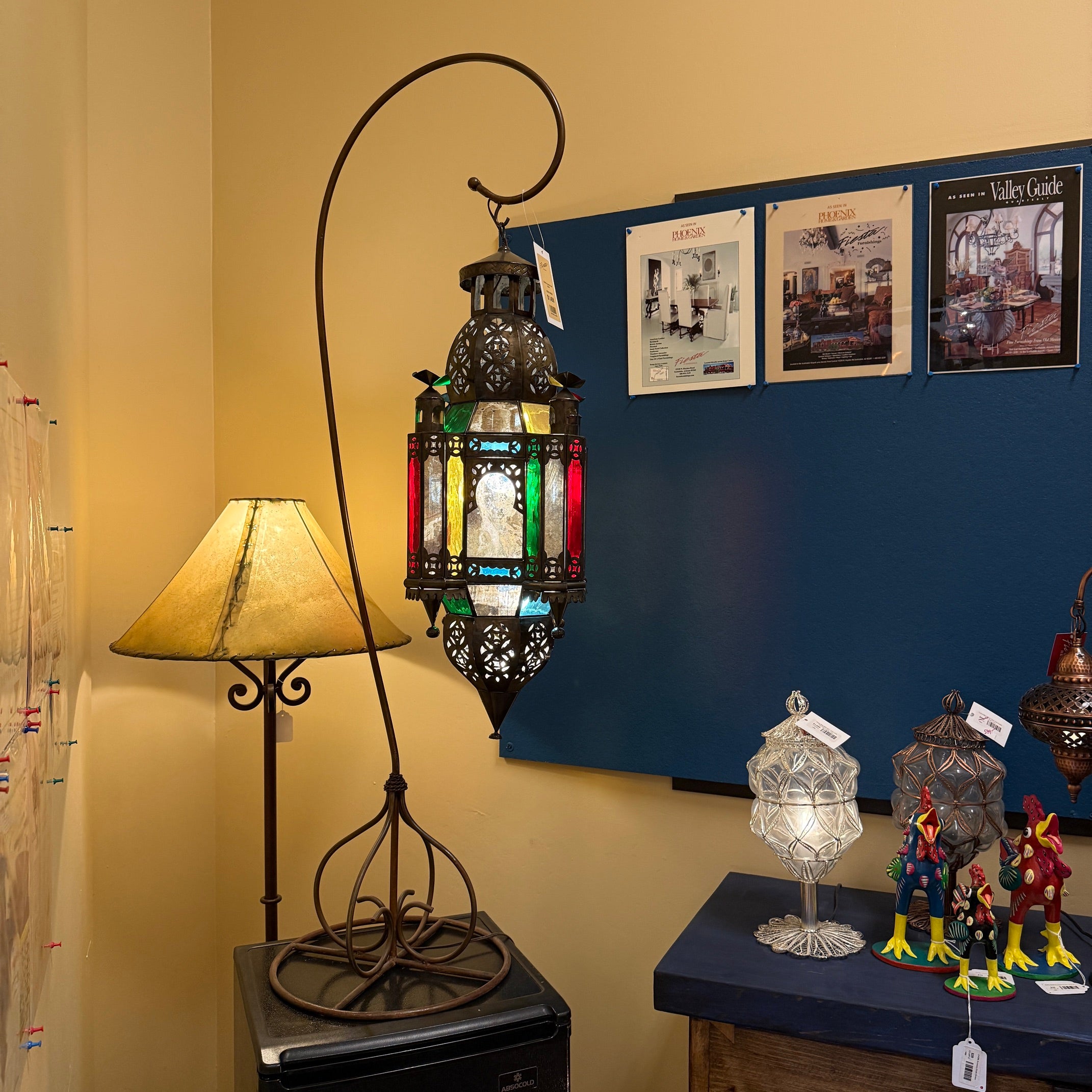 A decorative metal floor lamp with a tall curved stand and a hanging lantern featuring stained-glass-style panels in green, red, yellow, and blue, shown in a room setting next to another table lamp.