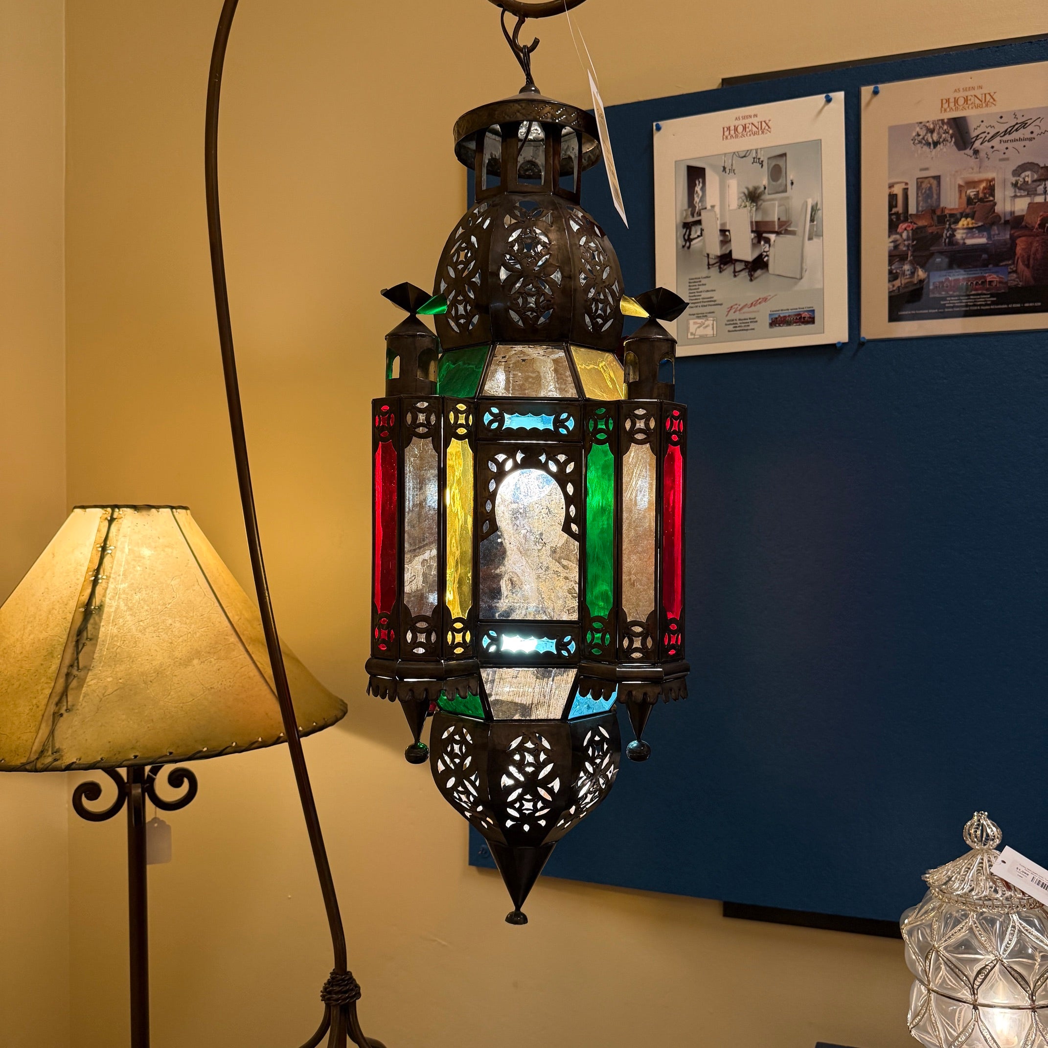 A hanging Moroccan-style lantern with intricate cutout metalwork and multicolored stained glass panels, shown in a room setting next to a floor lamp and against a blue wall.