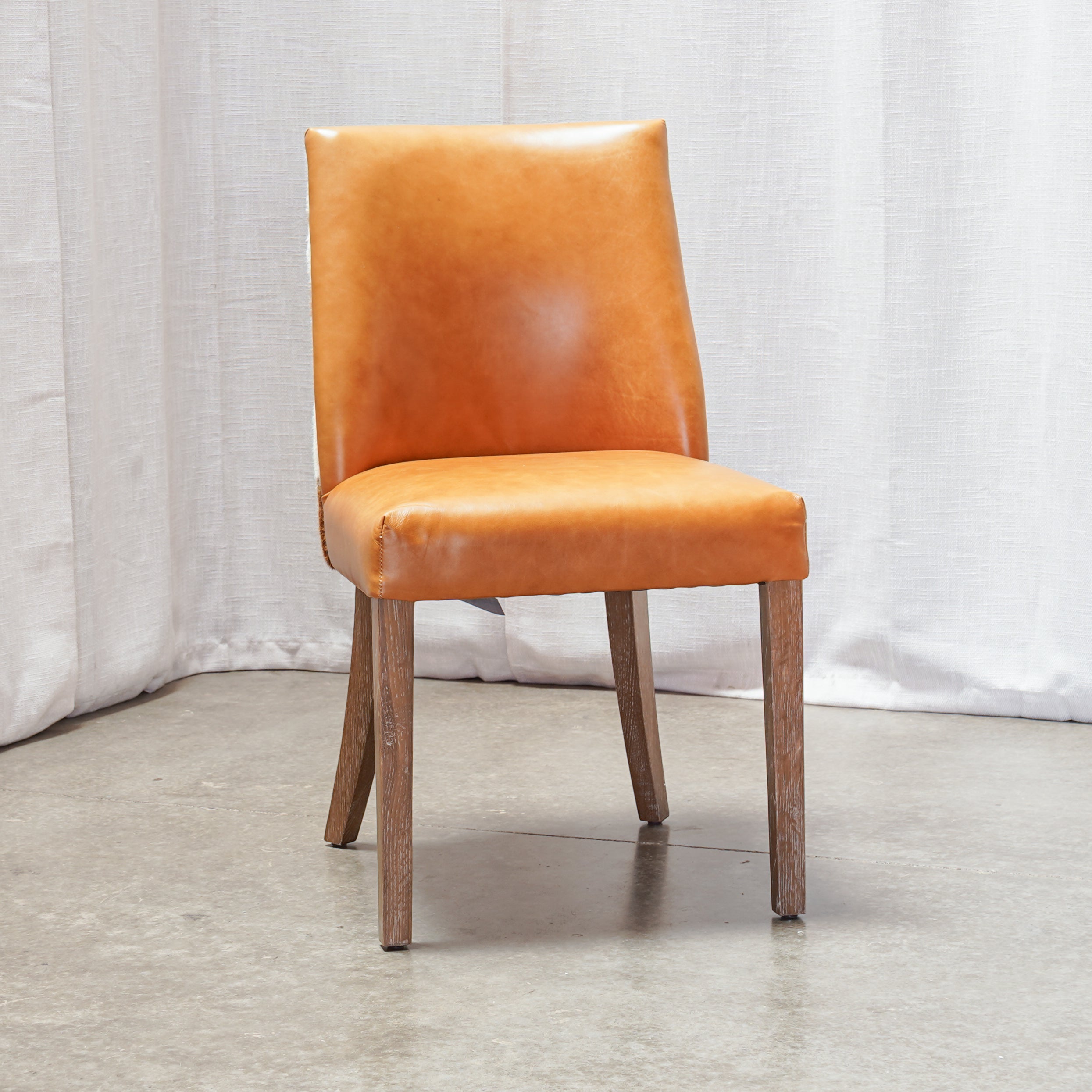An orange leather upholstered dining chair with a rectangular backrest and four tapered wooden legs, set against a white curtain backdrop on a concrete floor.