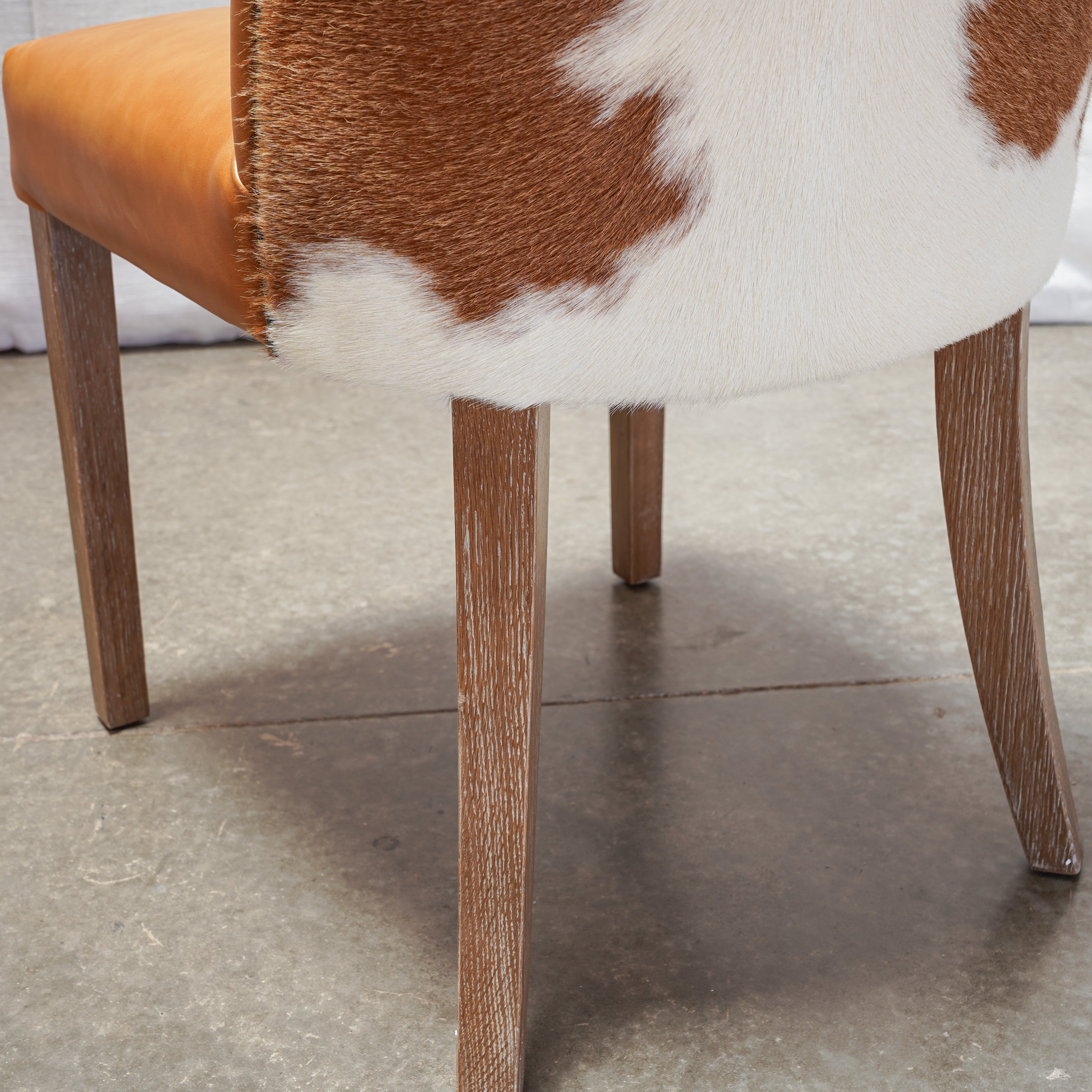 Close-up of a chair or stool with a brown and white cowhide upholstered seat and back, tan leather side panel, and medium brown wooden legs on a concrete floor.