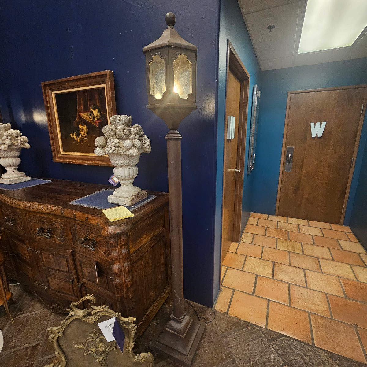 A tall dark metal and glass street lantern stands on a stone floor next to a dark wood cabinet in a hallway with deep blue walls. The lantern is lit, casting a warm glow, and a framed picture and white decorative urns sit on the cabinet.