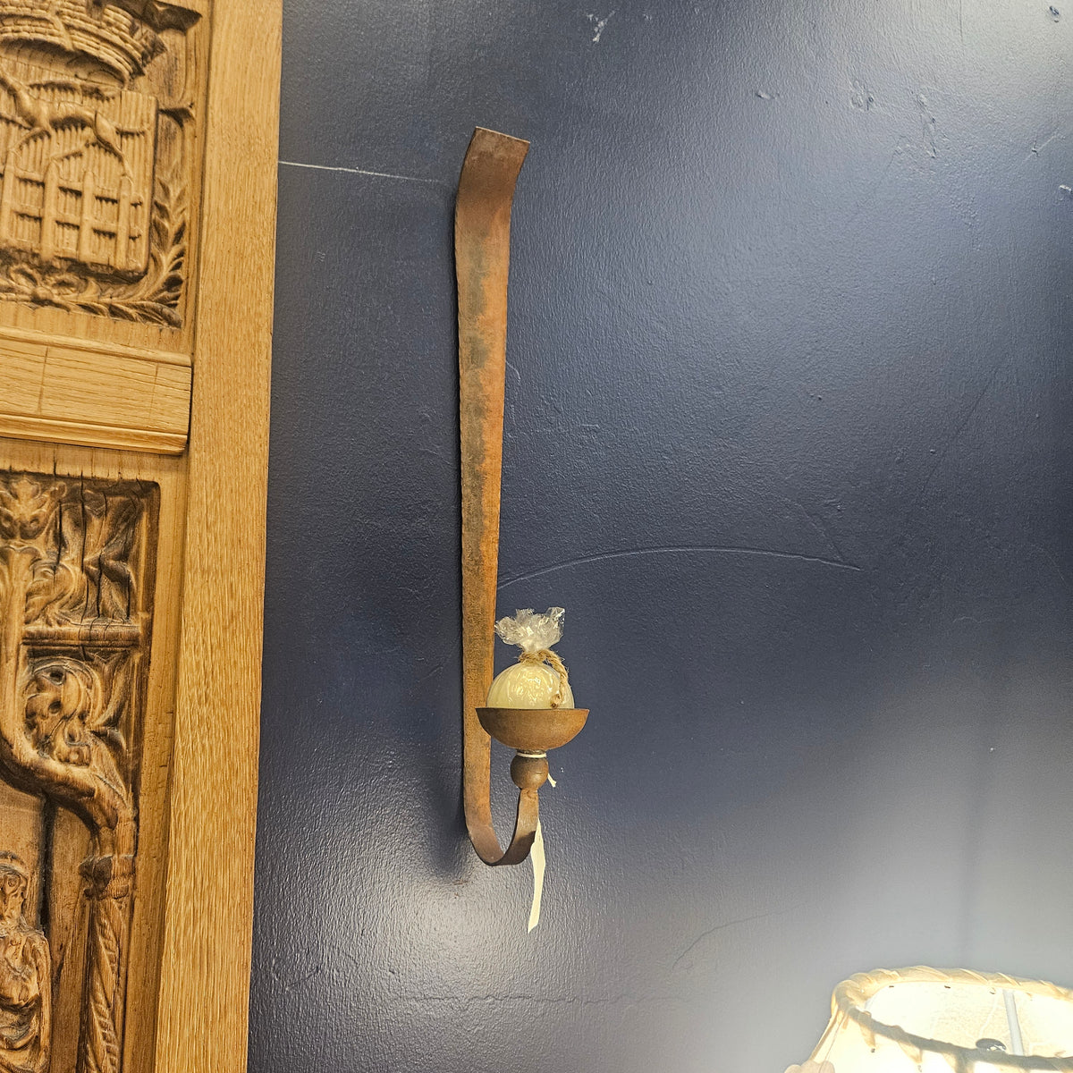 A rustic, weathered metal wall sconce with a curved hook design and a small glass cup holder, mounted on a dark blue-gray wall next to a carved wooden panel.