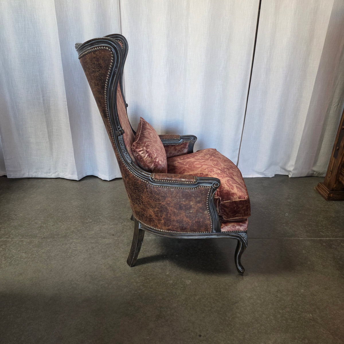 A distressed brown wingback armchair with a matching throw pillow, featuring dark carved wood trim and nailhead details, set against a white curtain backdrop.